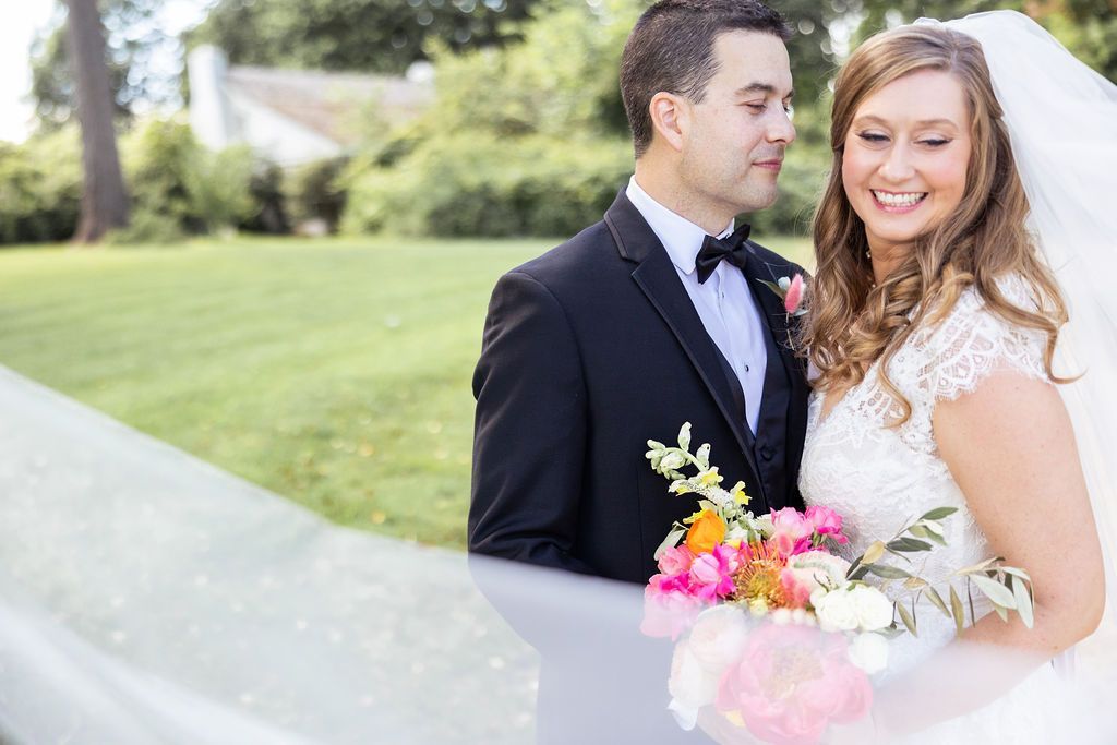 A bride and groom pose outdoors, smiling as the bride holds a vibrant, colorful bouquet with a veil draping in front.