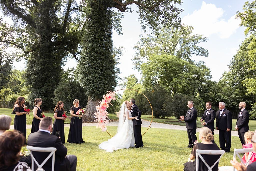 A bride and groom kiss under a floral arch during an outdoor wedding ceremony with a wedding party and guests nearby.