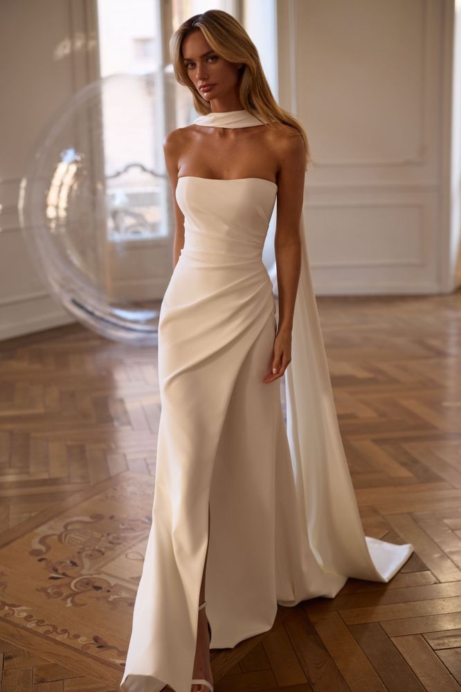 A model wears a strapless, white, floor-length wedding gown with a draped skirt and a matching neck scarf in a bright room.