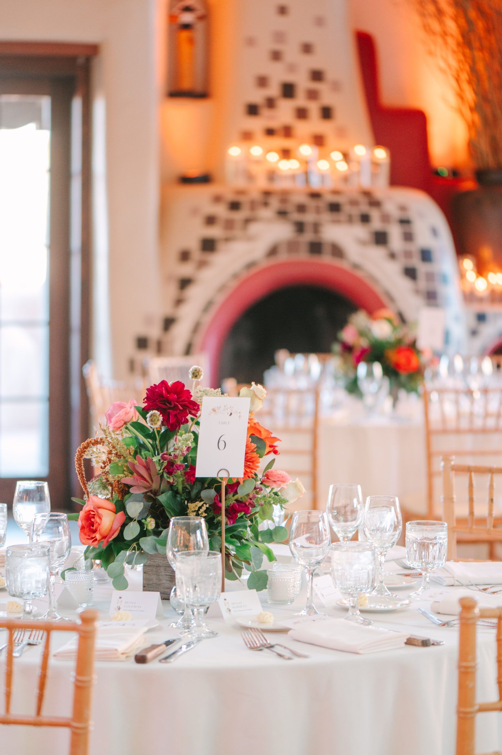 A table setting with a floral centerpiece and number 6 sign, in front of a fireplace in a softly lit event space.