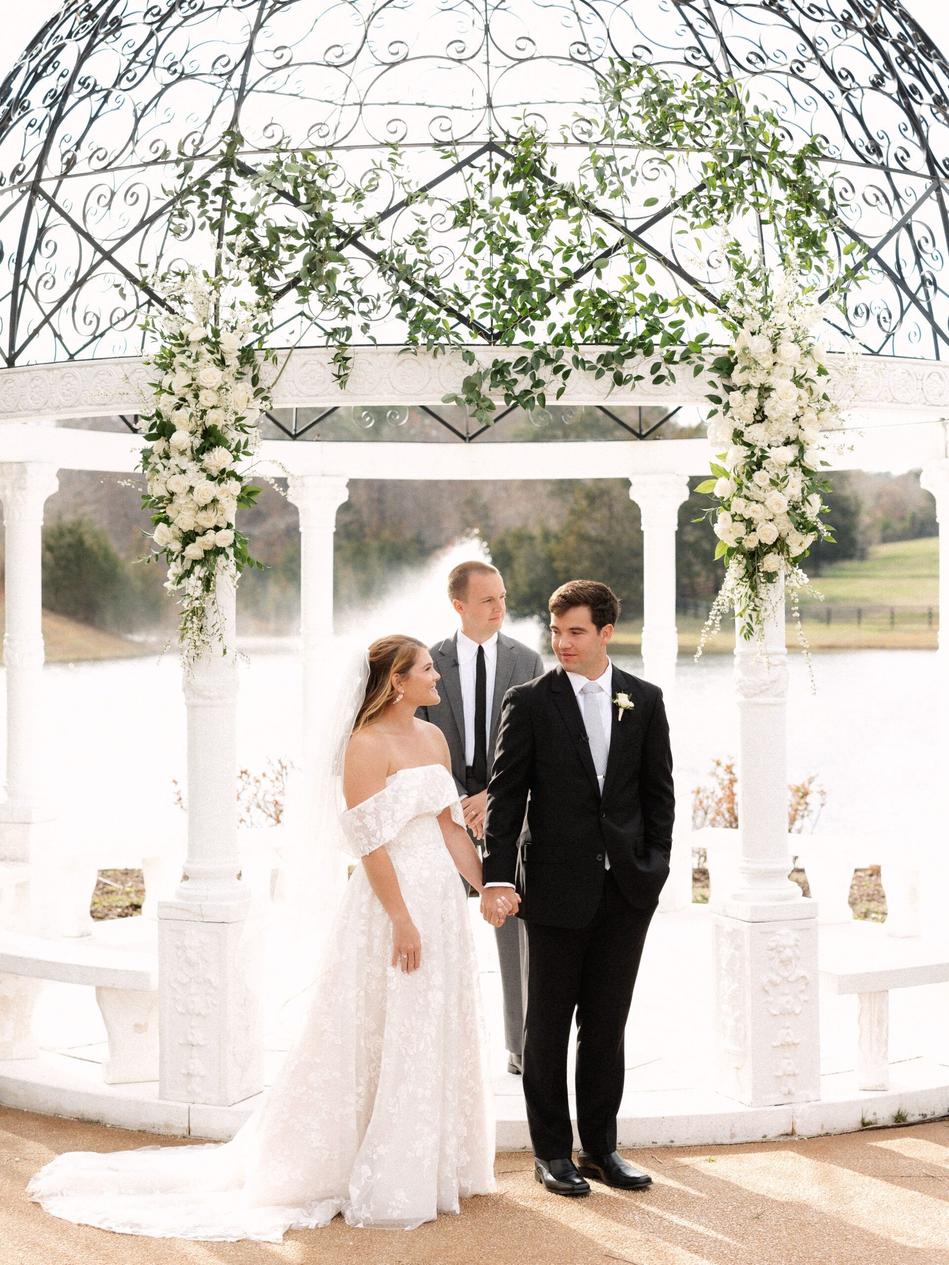 A bride and groom hold hands under a white floral gazebo by a lake, standing before an officiant during their ceremony.