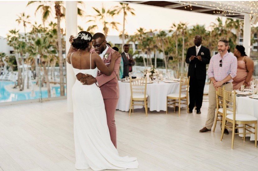 A couple in wedding attire shares a dance on a terrace by a pool, observed by guests seated at outdoor tables.