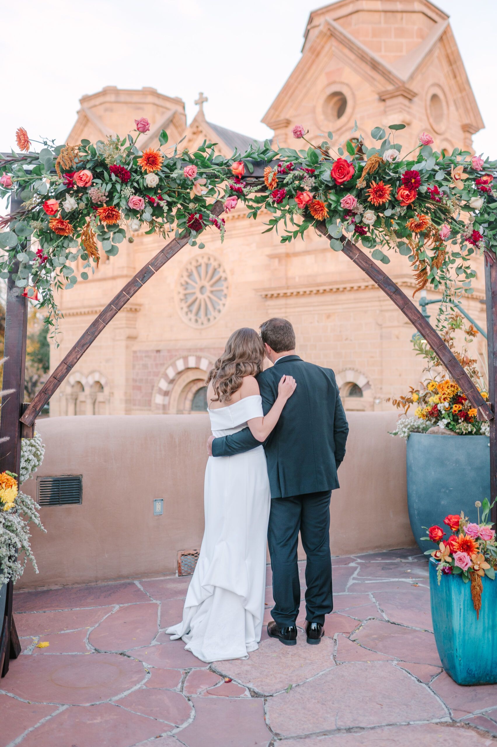 A couple in wedding attire stands under a flower-adorned arch, facing a historic adobe church with a stone courtyard.