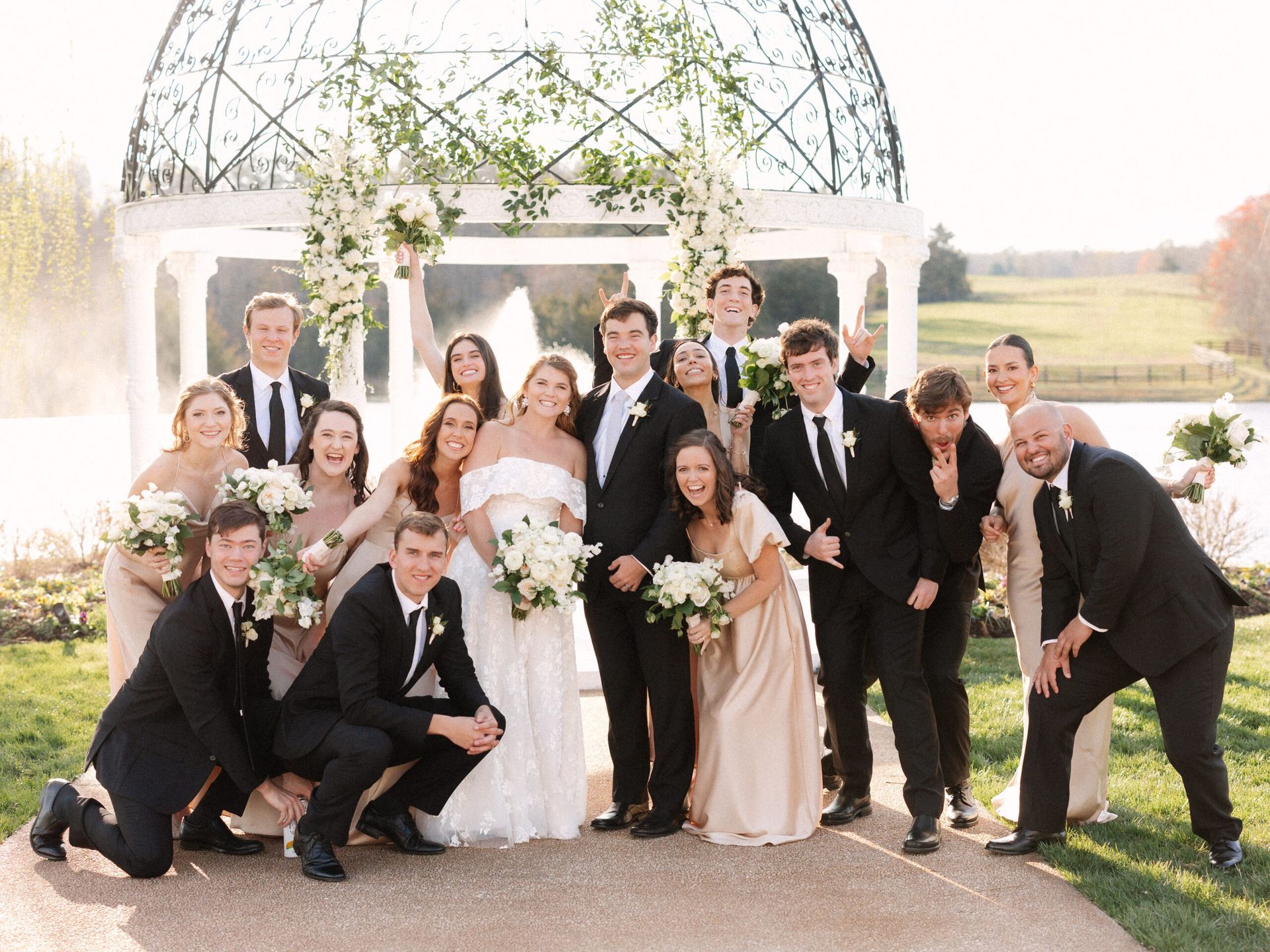 Wedding party poses in front of a floral gazebo by a lake, with some members kneeling and others standing.