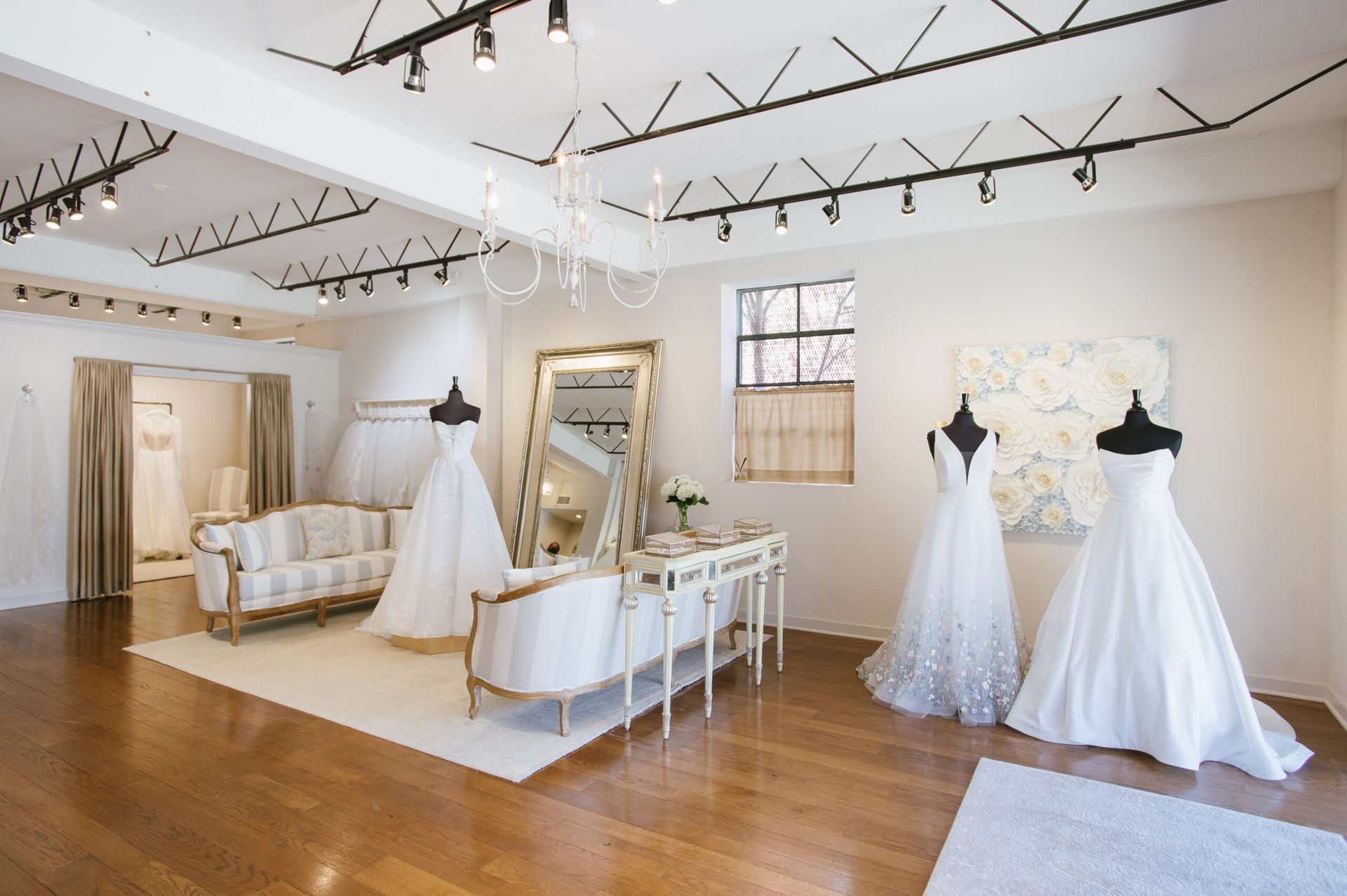 Bridal boutique interior with wedding dresses on mannequins, elegant seating, and a large mirror in a bright room.