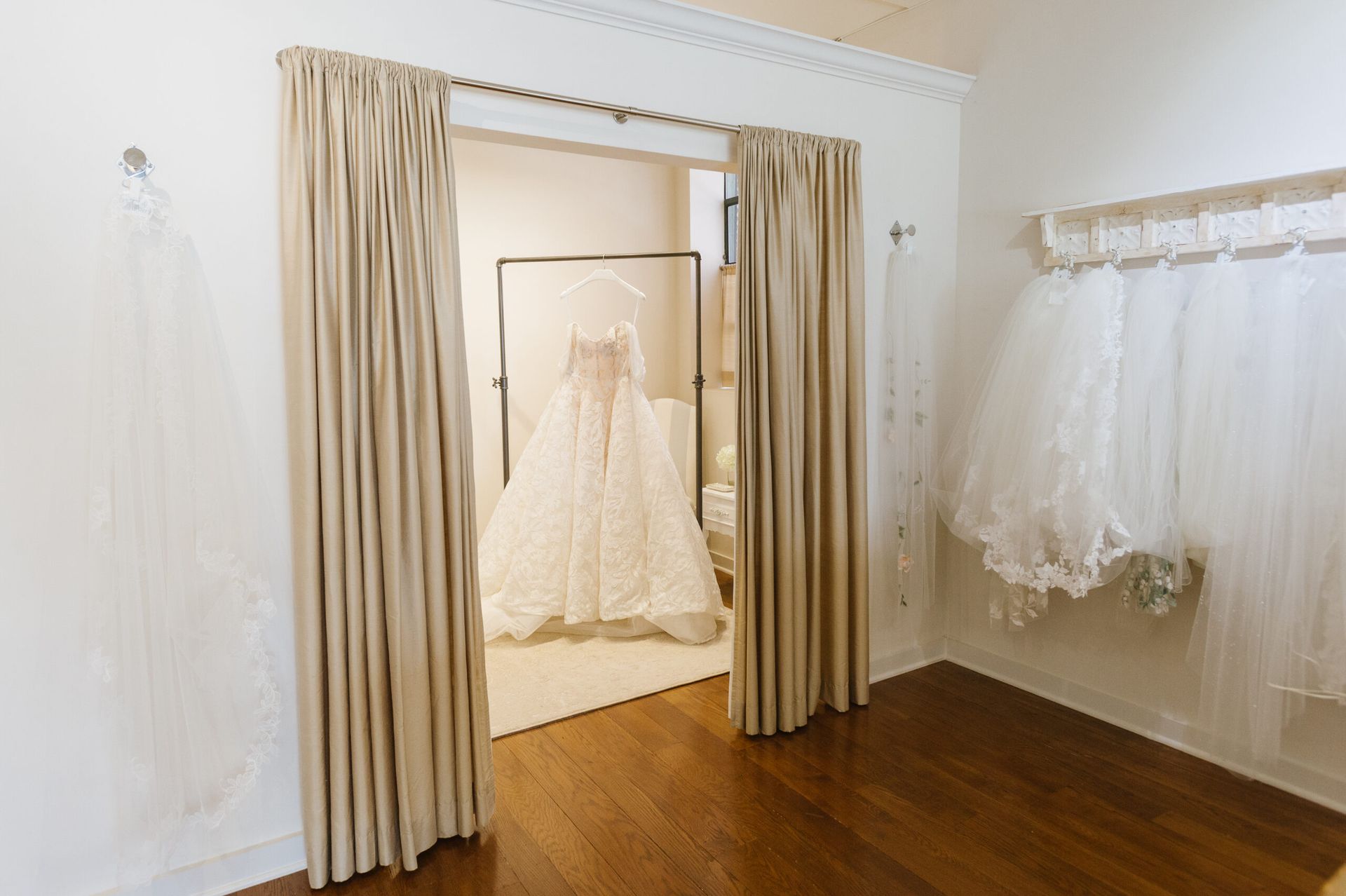 A wedding dress on a rack viewed through beige curtains, with white veils hanging on the wall of a bridal shop.