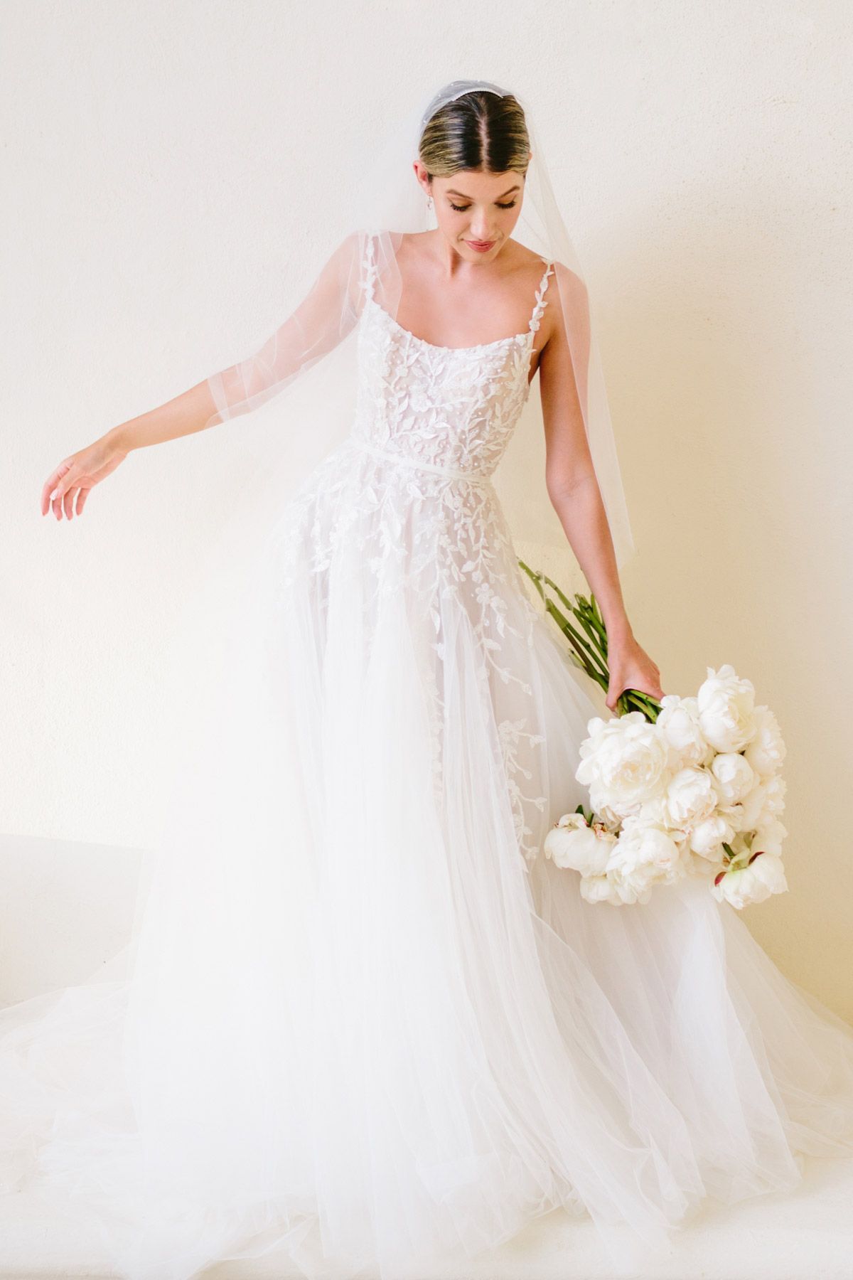 A person in a white wedding gown and veil holding a large bouquet of white flowers, looking down against a neutral wall.