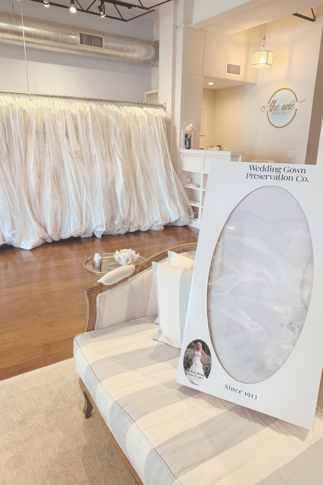 A striped settee with a wedding dress preservation kit in the foreground, with racks of bridal gowns in the background.