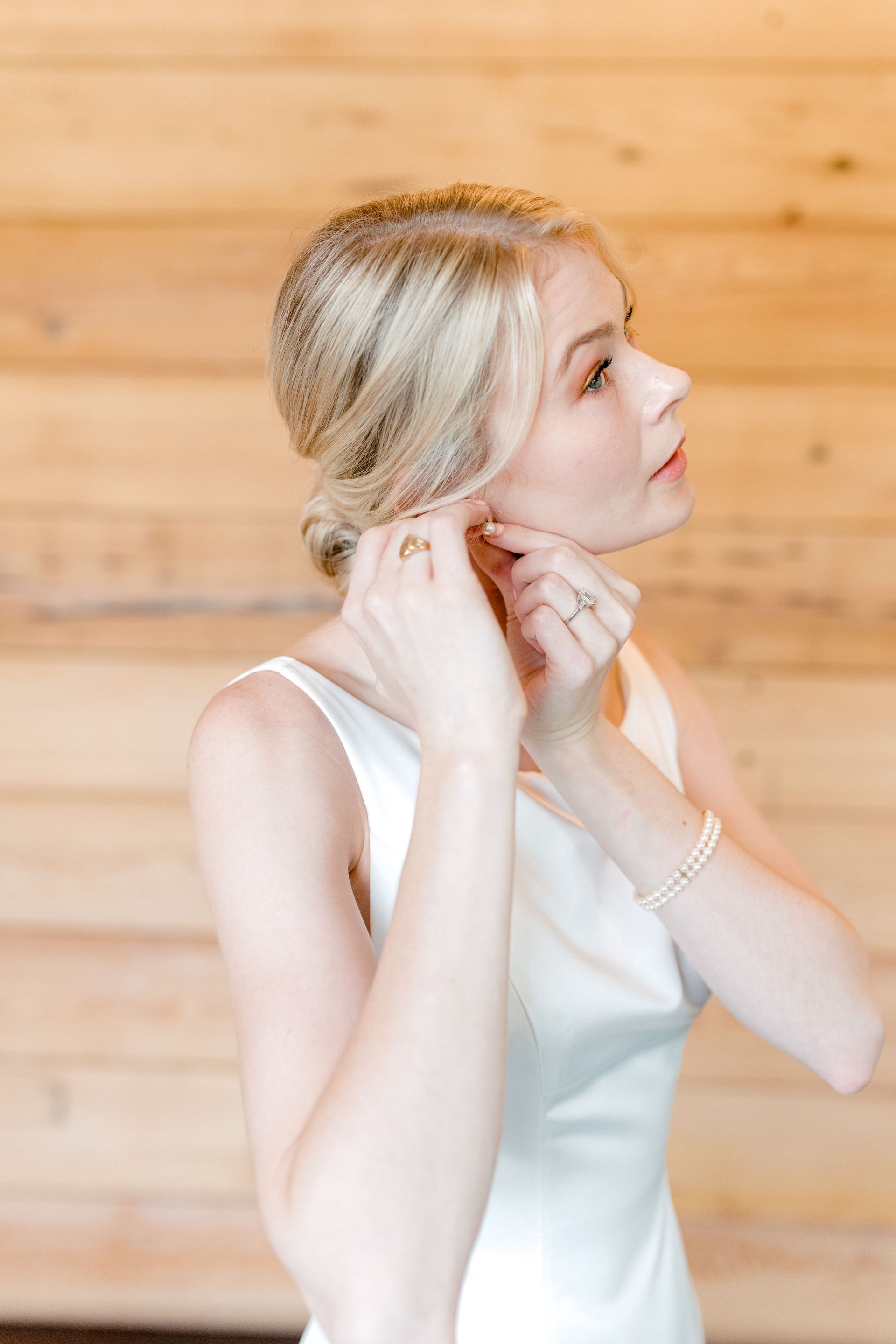 A person with blonde hair in an updo wears a white dress while putting on an earring against a rustic wood-plank wall.