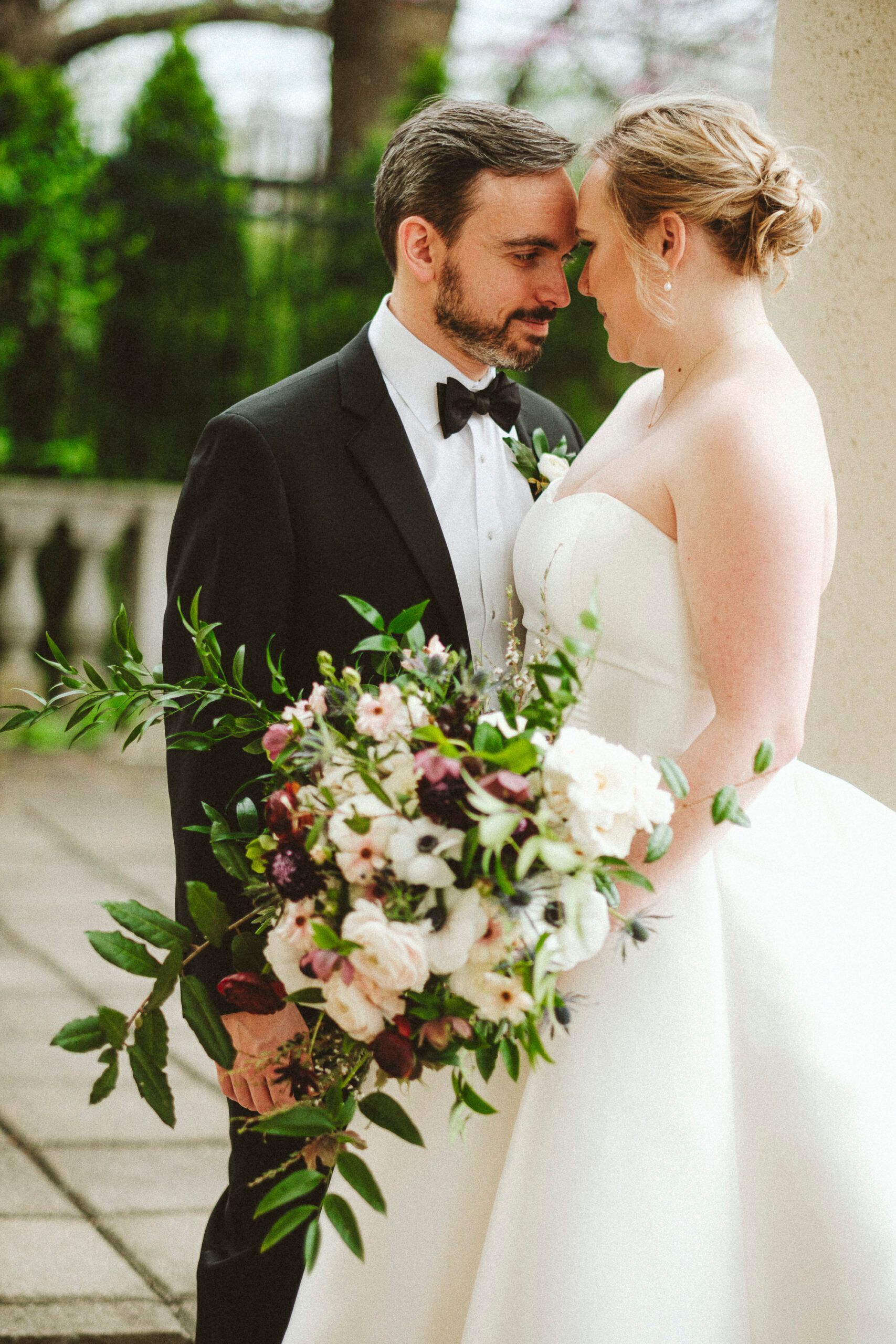 A couple in wedding attire faces each other while holding a lush floral bouquet on a stone patio.