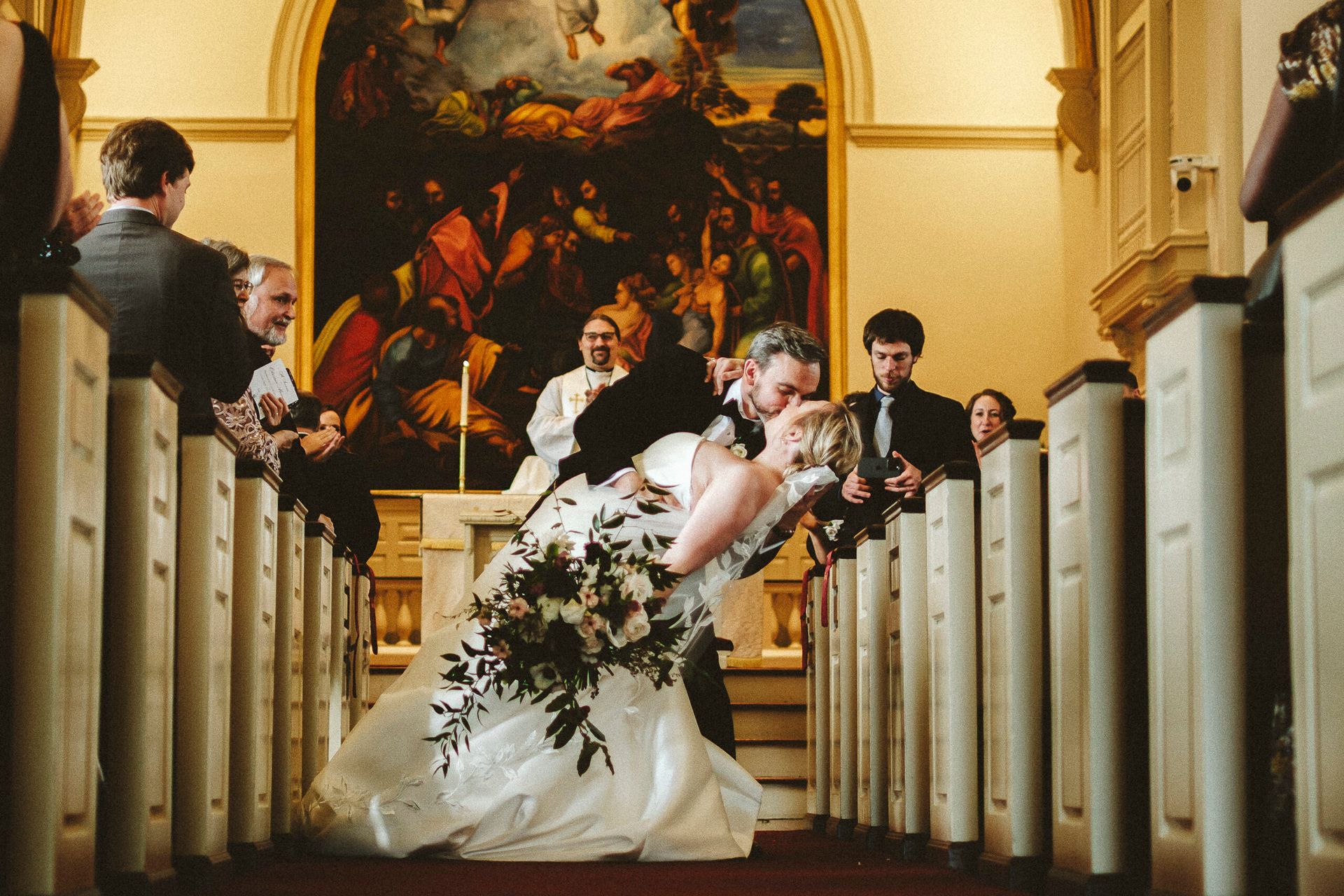 A couple in wedding attire kisses while dipping in the aisle of a church in front of an altar painting.