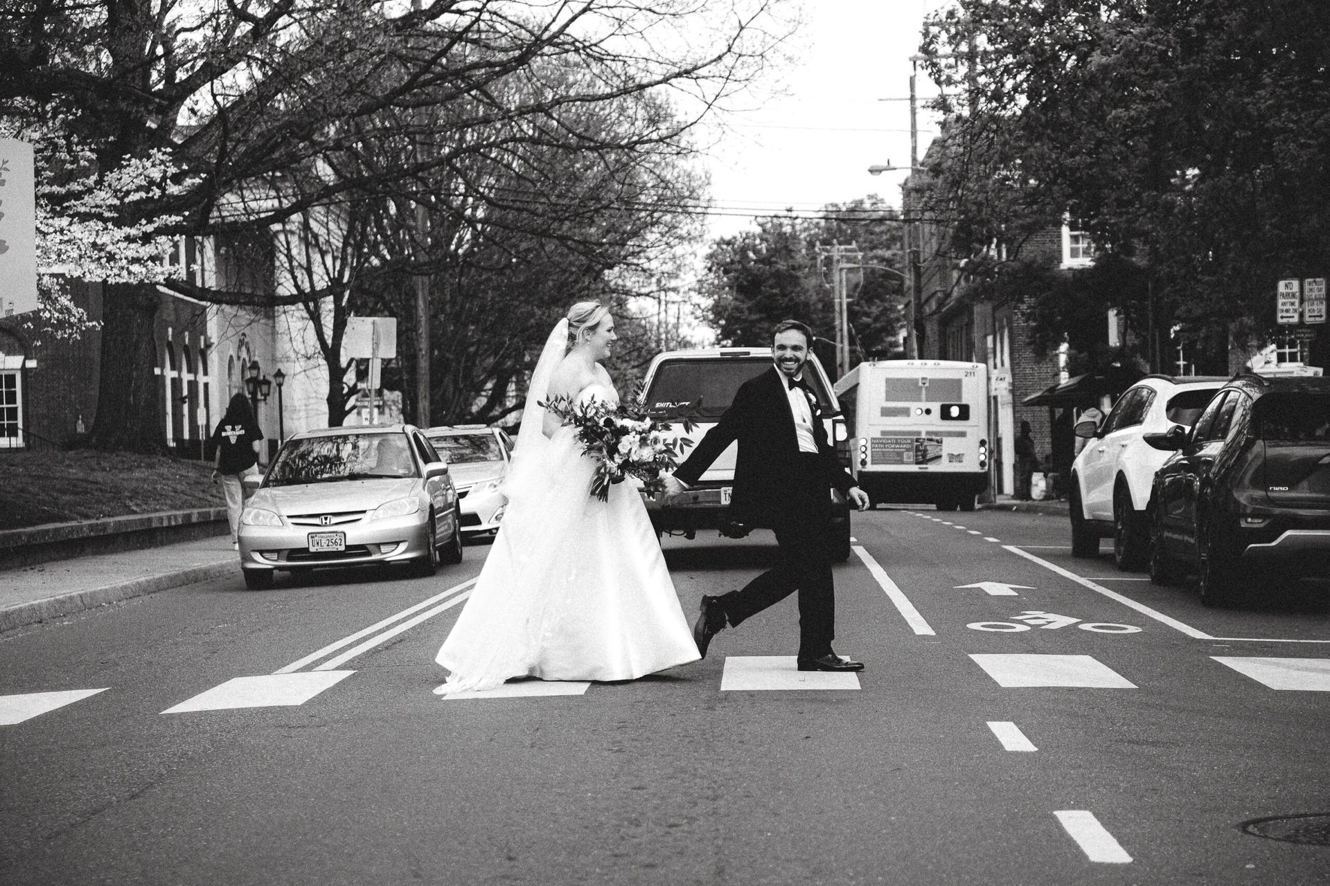 A bride in a white wedding gown and a groom in a tuxedo walk across a city street crosswalk, laughing together.