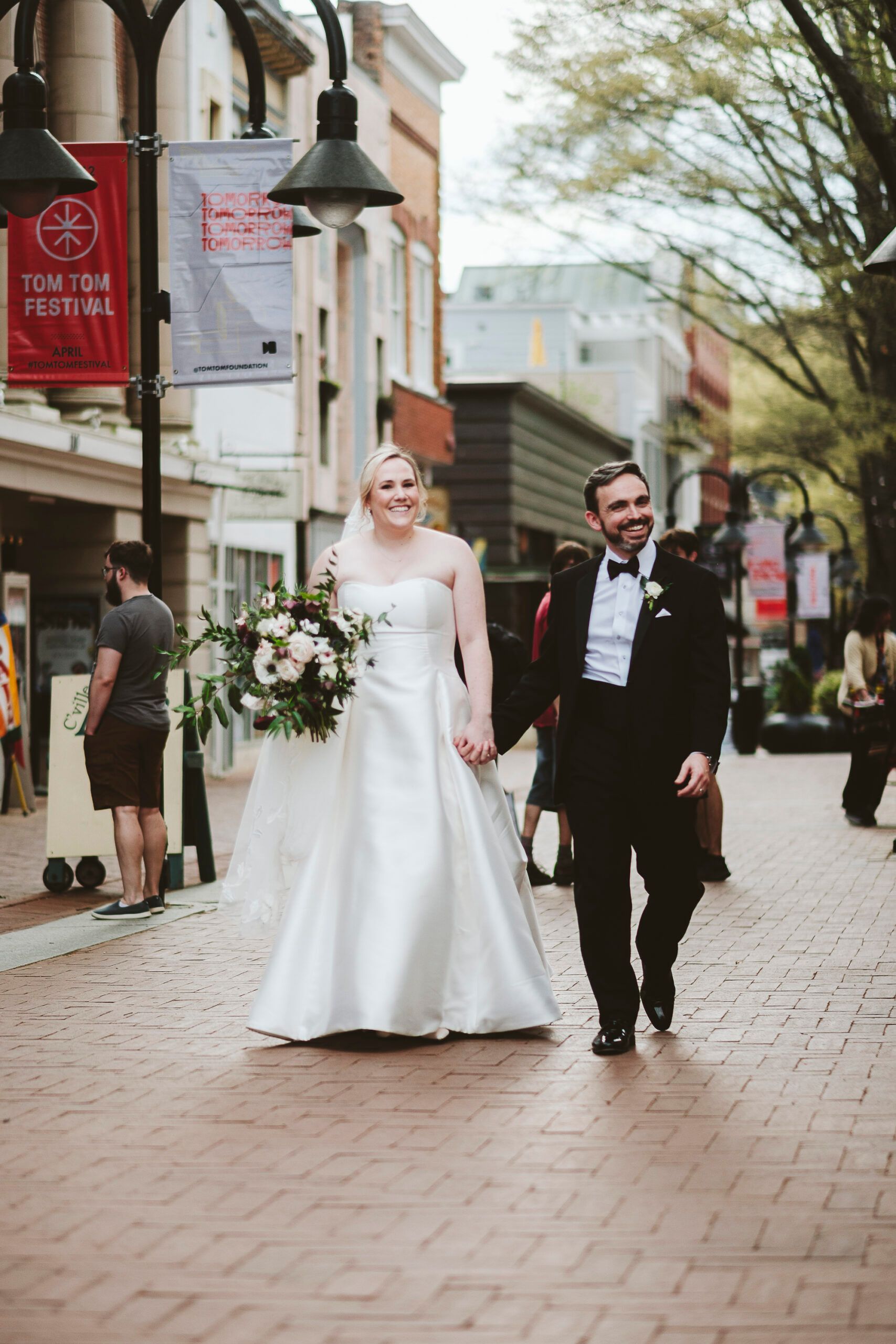 A smiling bride and groom in wedding attire walk hand-in-hand down a brick pedestrian street.