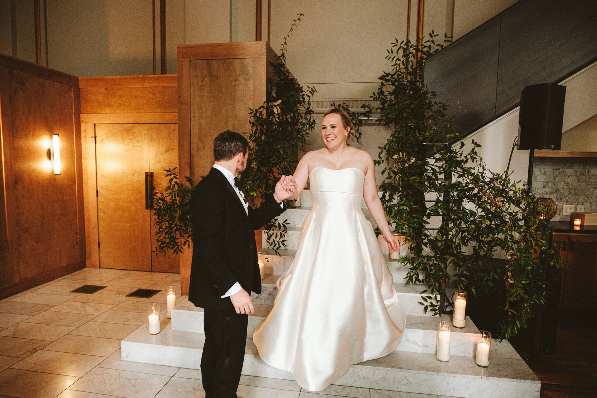 A bride in a white wedding gown walks down stone steps toward her partner in a tuxedo in a room decorated with greenery.