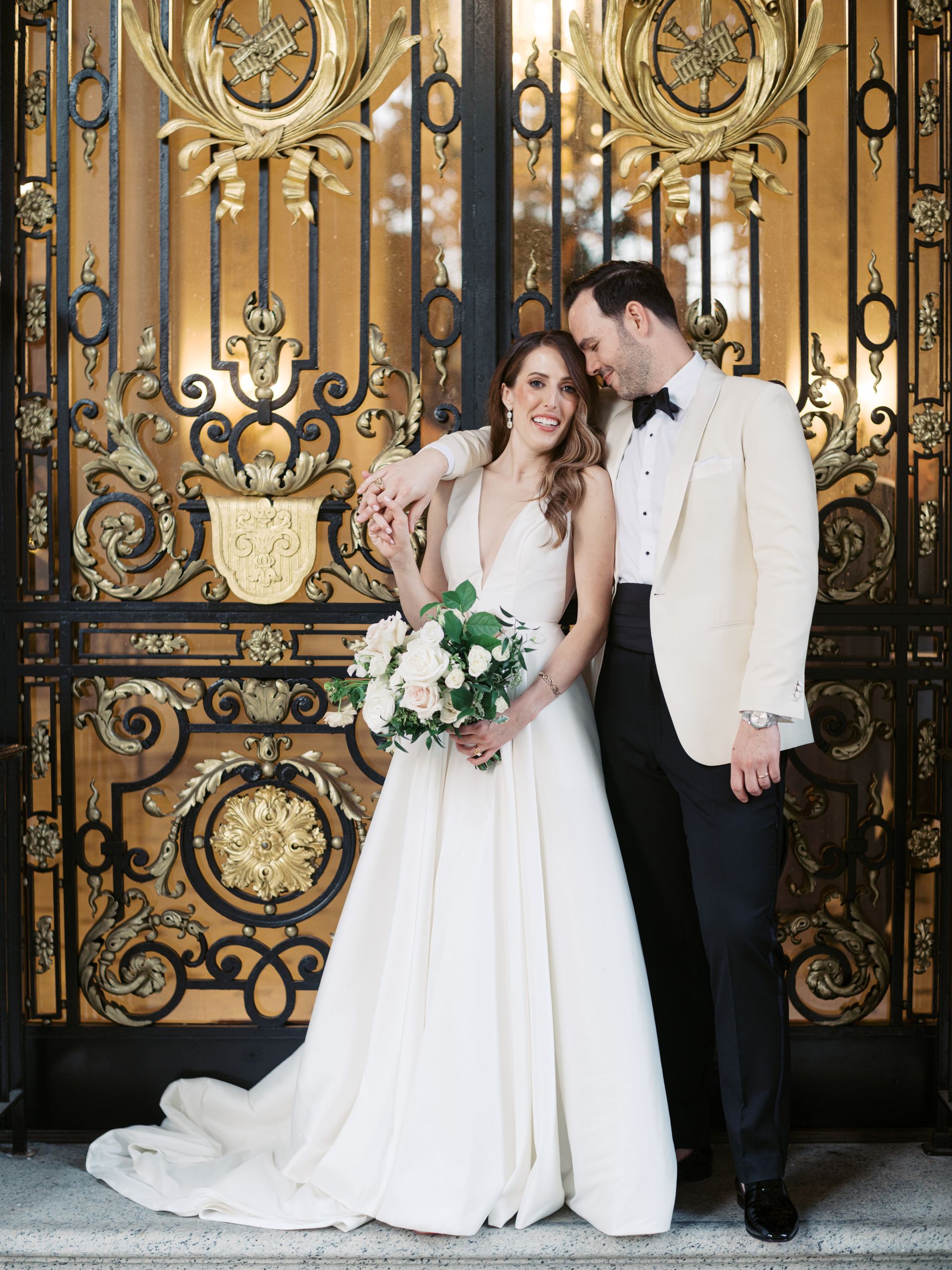 A bride in a white wedding gown and a groom in a tuxedo pose together in front of ornate, gold-accented metal gates.