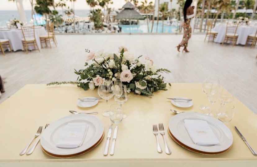 A table set for two at an outdoor wedding reception overlooking the ocean, featuring white flowers and gold-trimmed plates.