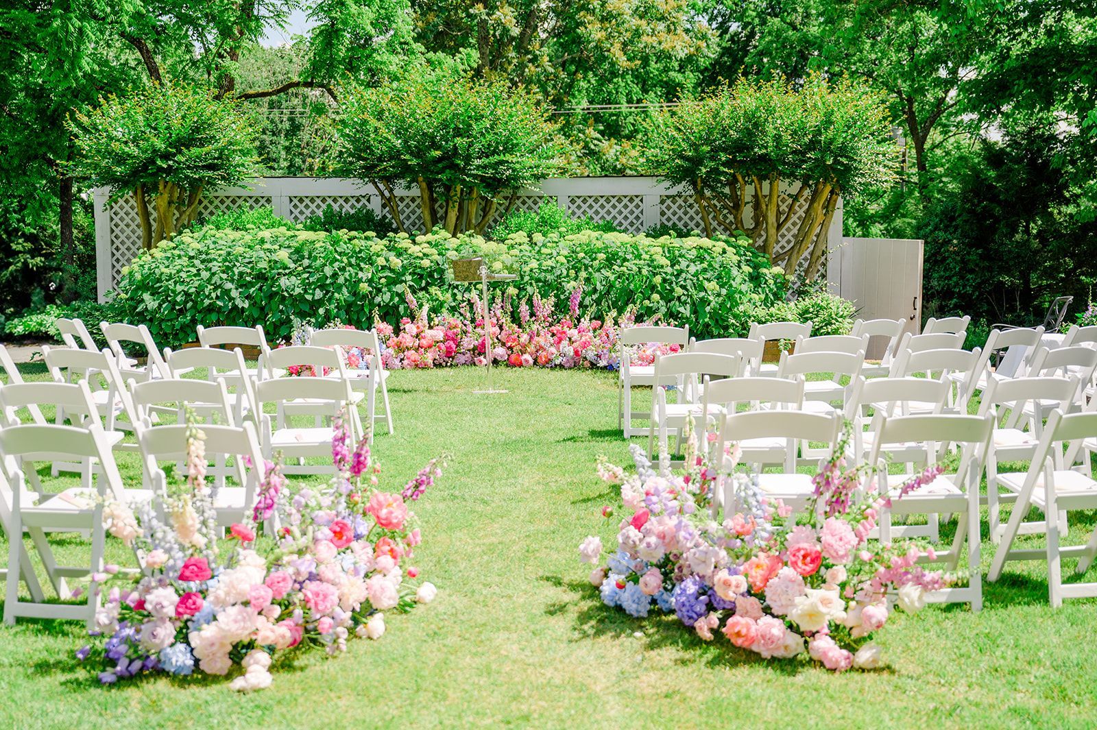 Rows of white chairs face an outdoor wedding aisle adorned with lush, colorful floral arrangements on a grassy lawn.