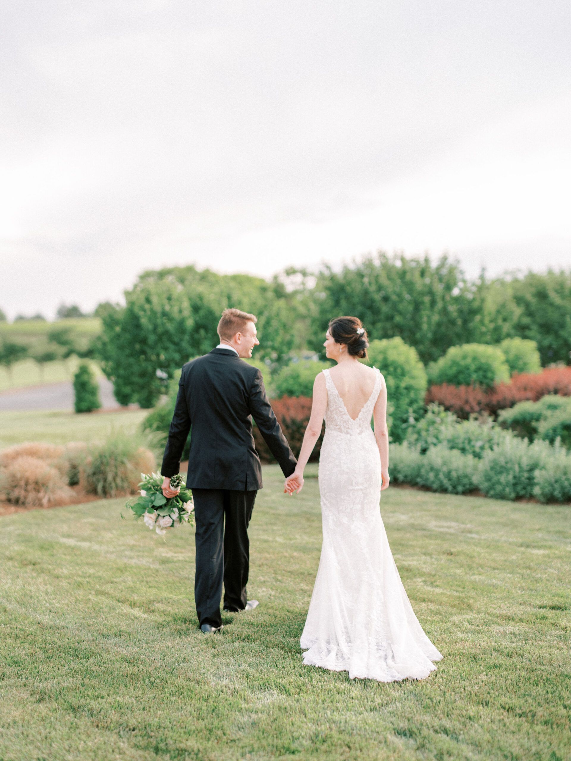 A couple in wedding attire walks hand-in-hand away from the camera through a grassy field with trees in the background.