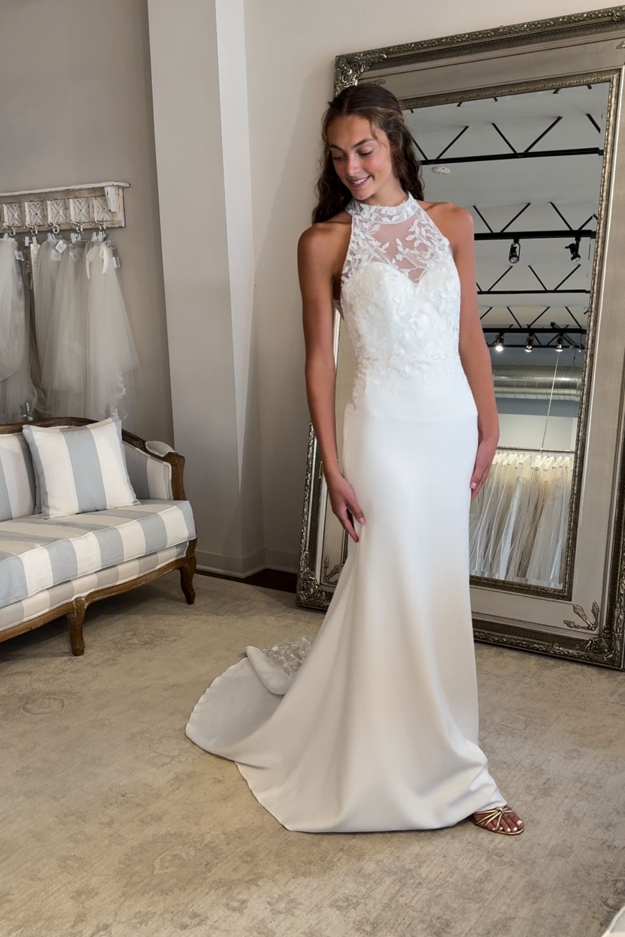 A person wearing a white, floor-length wedding dress with a high-neck lace bodice, standing in a bridal boutique.