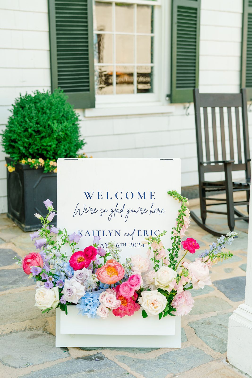 A white wedding welcome sign for Kailyn and Eric, decorated with vibrant pink, white, and blue flowers, on a stone patio.