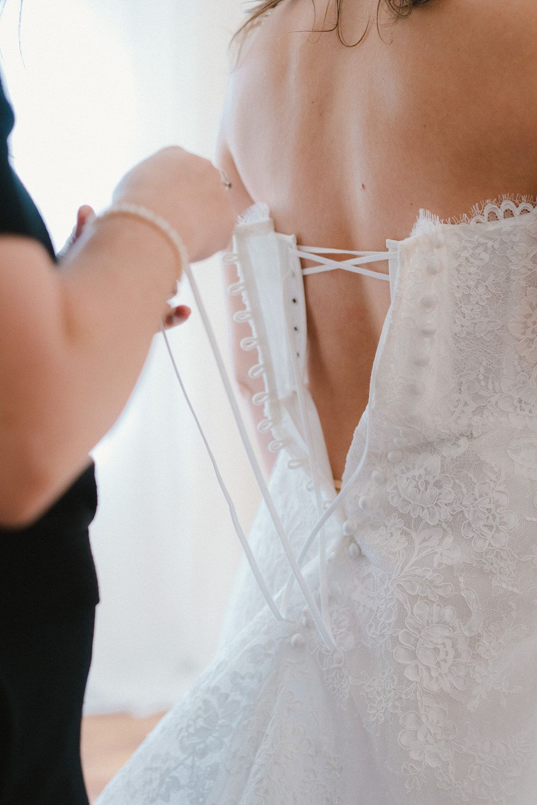 A person in a black shirt laces a white lace bridal gown on someone's back.