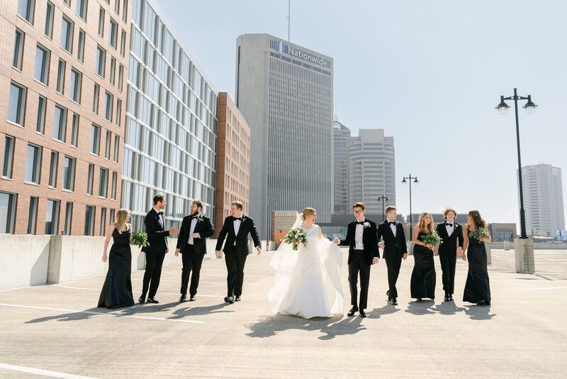 A wedding party walks across a rooftop parking deck with modern skyscrapers in the background on a sunny day.