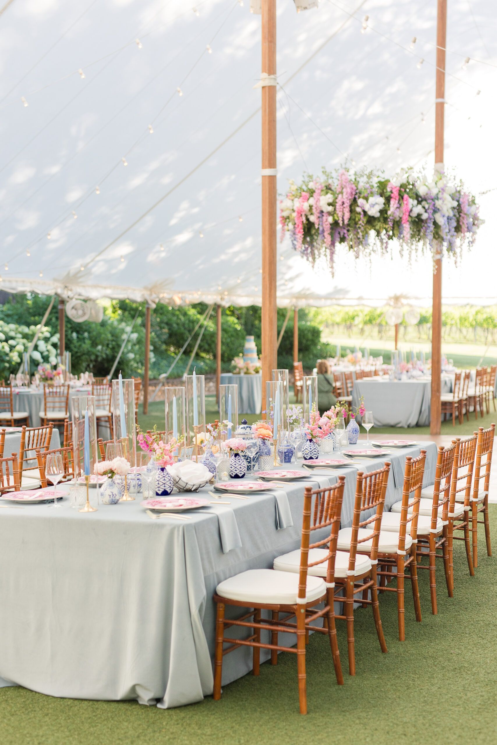 A long banquet table set for a wedding under a white tent, featuring pastel floral arrangements and wooden cross-back chairs.