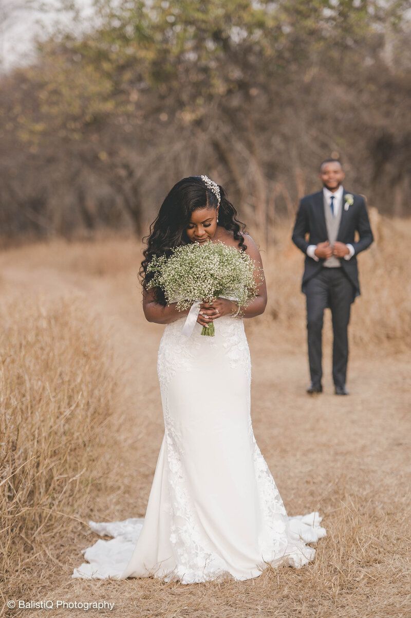 A person in a white wedding gown holding a bouquet stands in a field, with a person in a suit standing in the background.