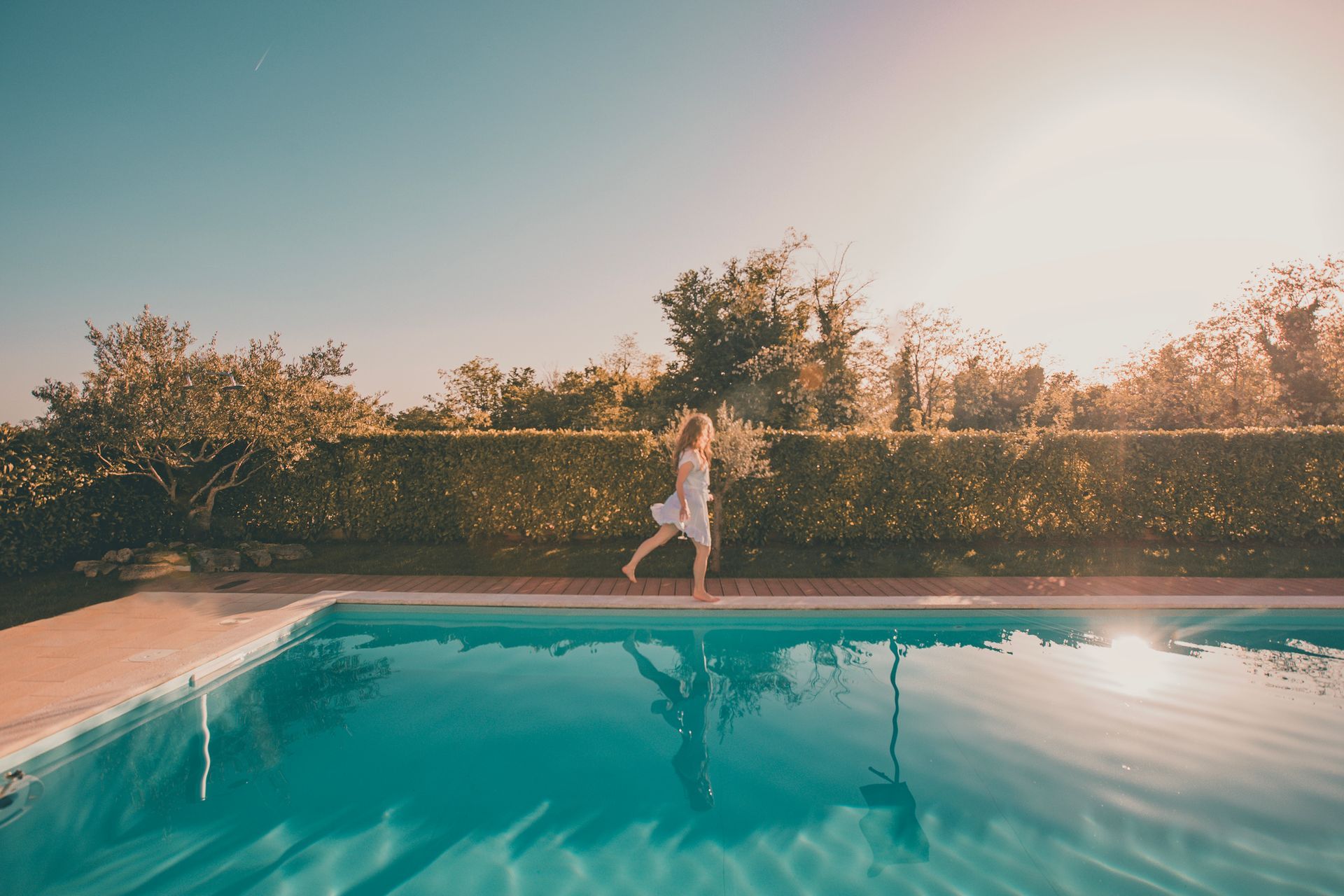 Woman in white dress runs alongside a pool, sun shining brightly, trees and blue sky.