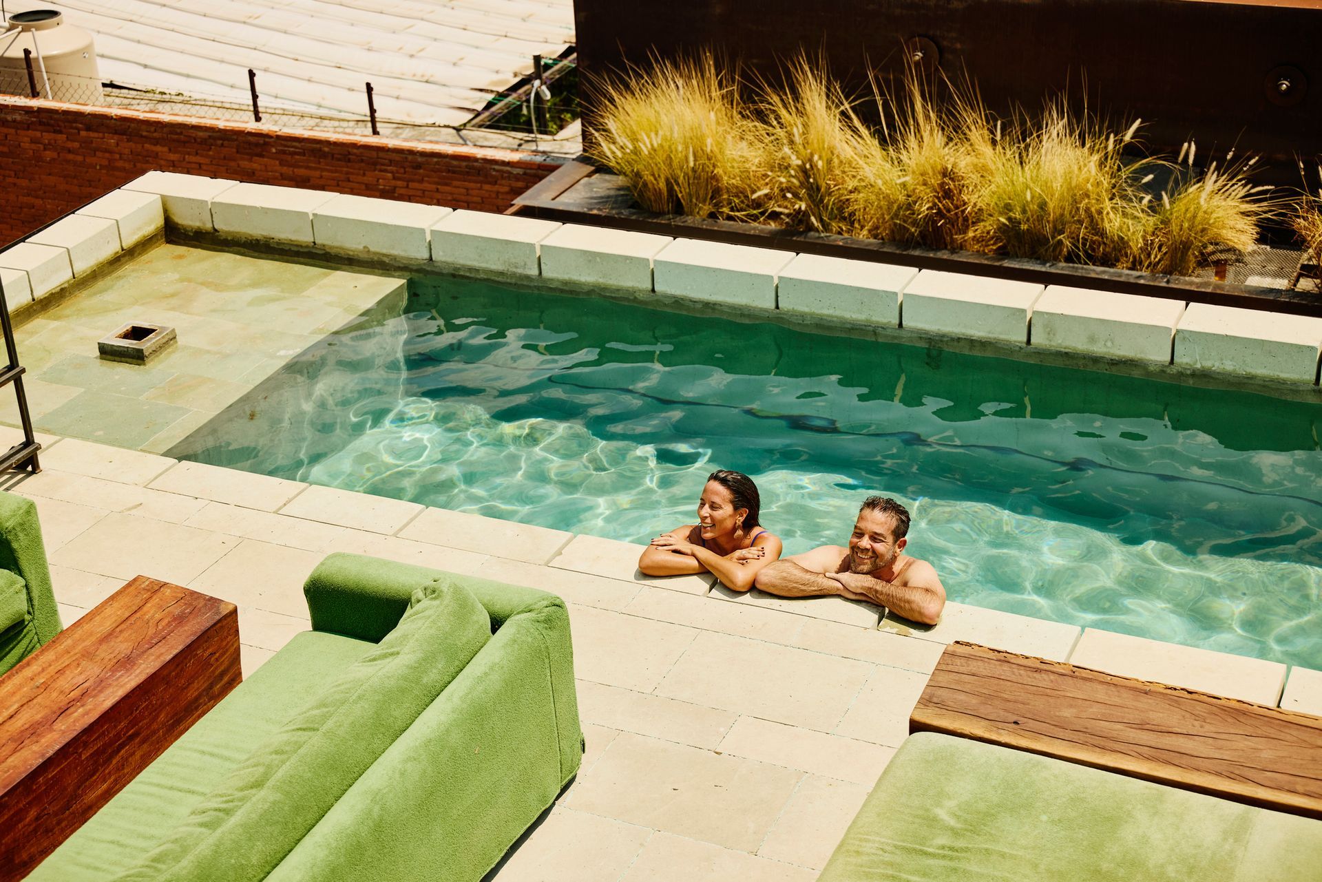 Two people smiling in a rooftop pool, green and tan decor, sunny.