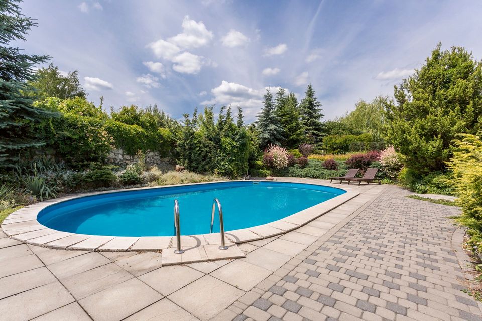 Oval swimming pool with blue water, surrounded by a stone patio and greenery under a blue sky with clouds.