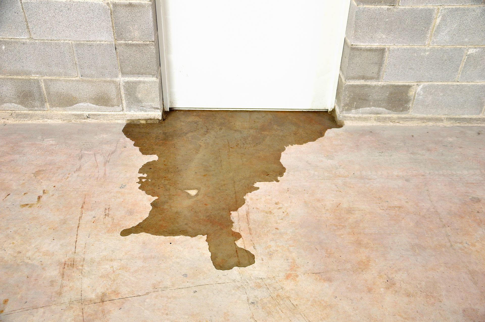 Wet stain on a concrete floor below a white door and cinder block wall