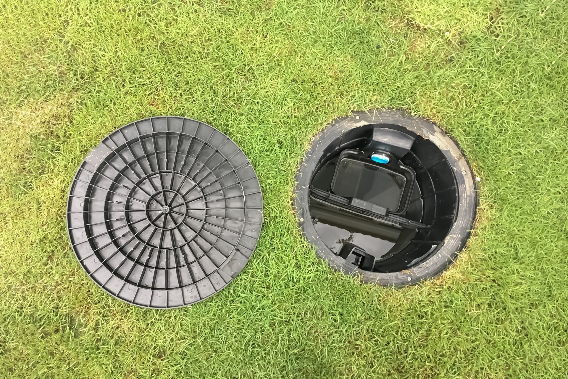Open manhole in grass with a round black cover beside it