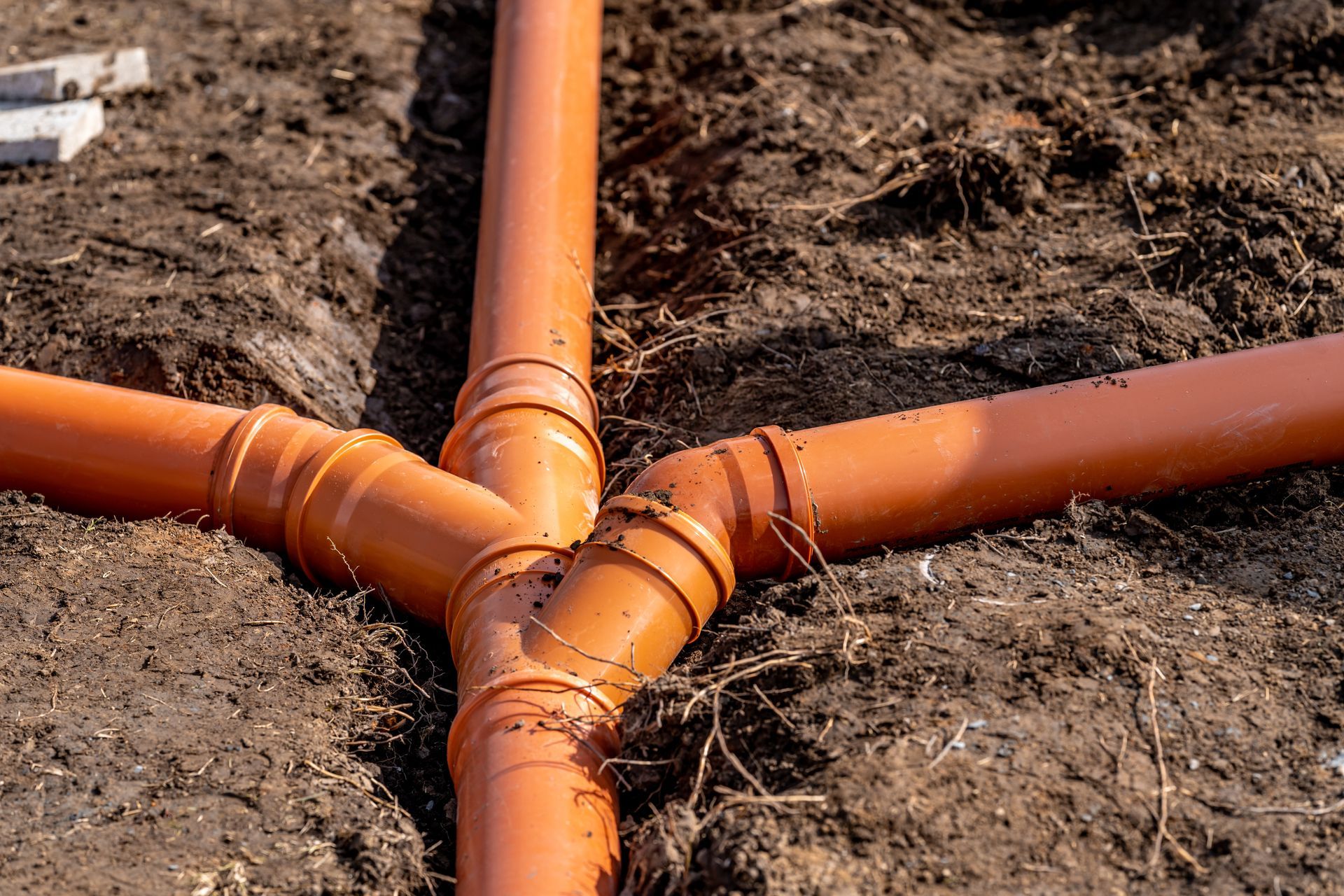 Orange drainage pipes joining in a Y-shape beneath soil in a trench.