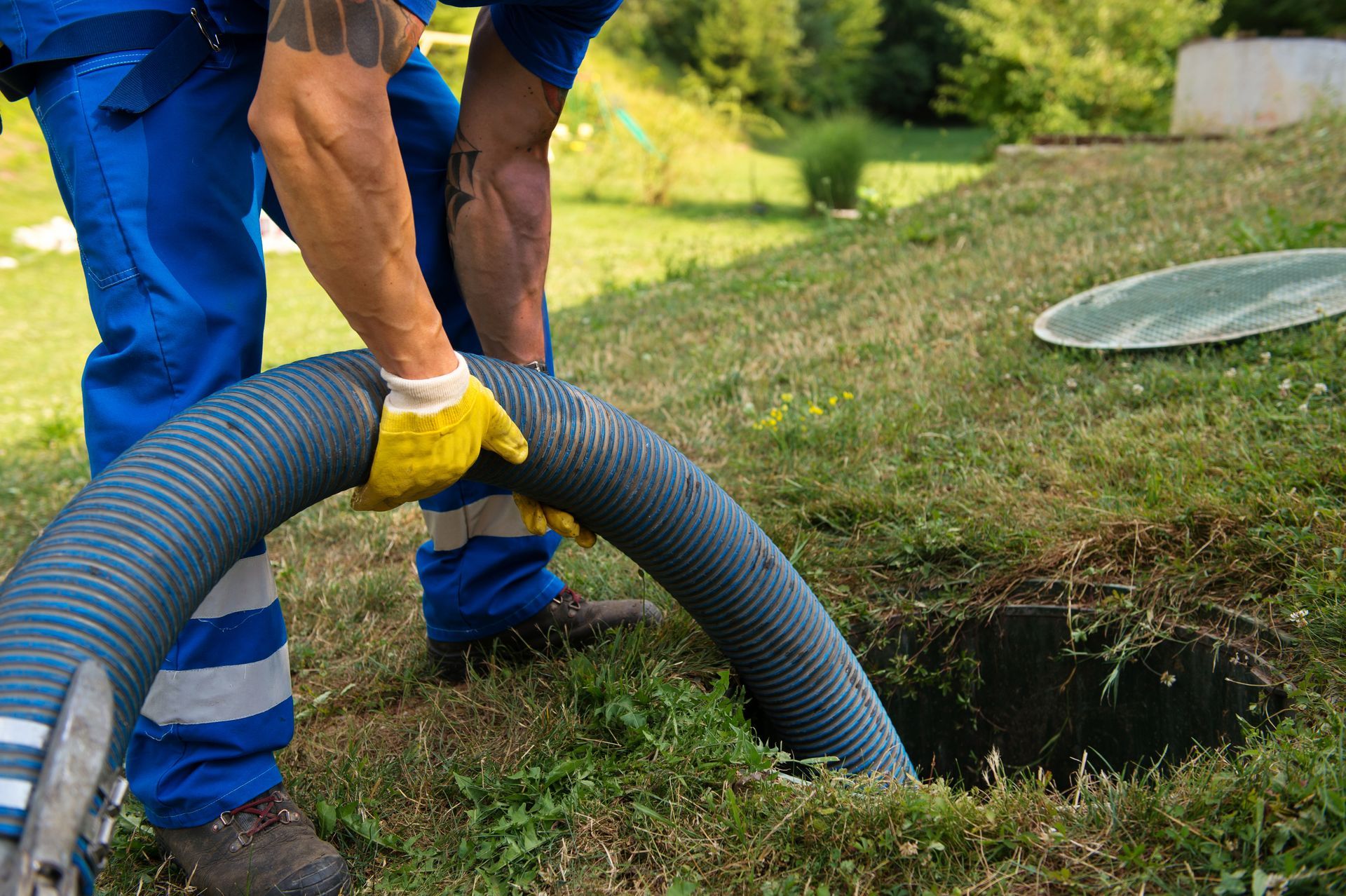 Worker in blue overalls handling a gray hose beside an open manhole on grass.