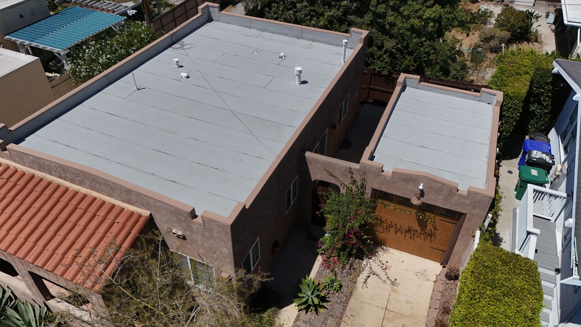 An aerial view of a two-story brown stucco house with a flat roof, garage, and green landscaping.