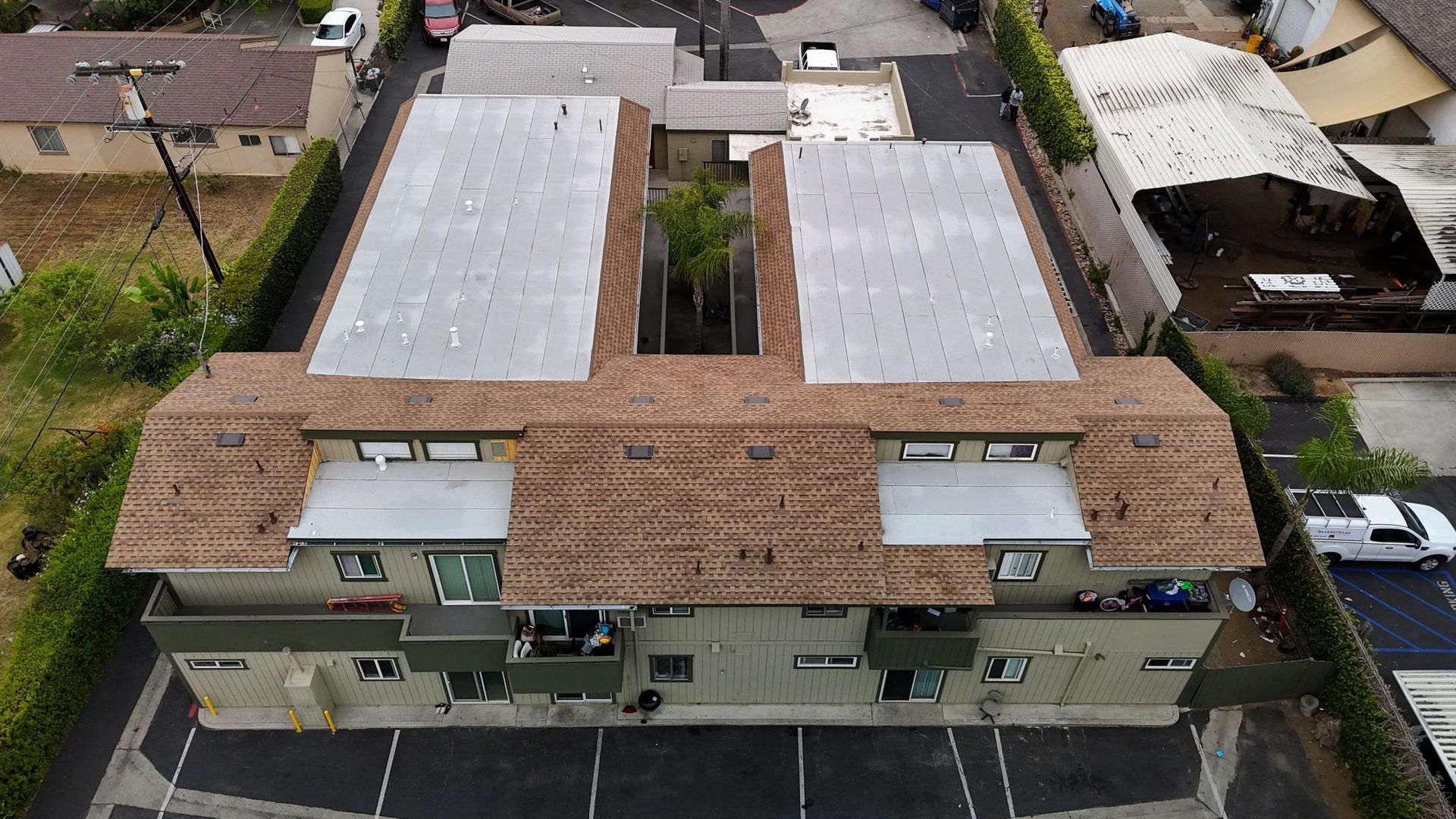 An aerial view of a two-story apartment building with a brown roof and green exterior, surrounded by trees and parked cars.