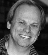 A black and white photo of a smiling man wearing a striped shirt.