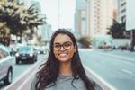 Woman with glasses smiles, standing on a city street with cars and buildings in the background.