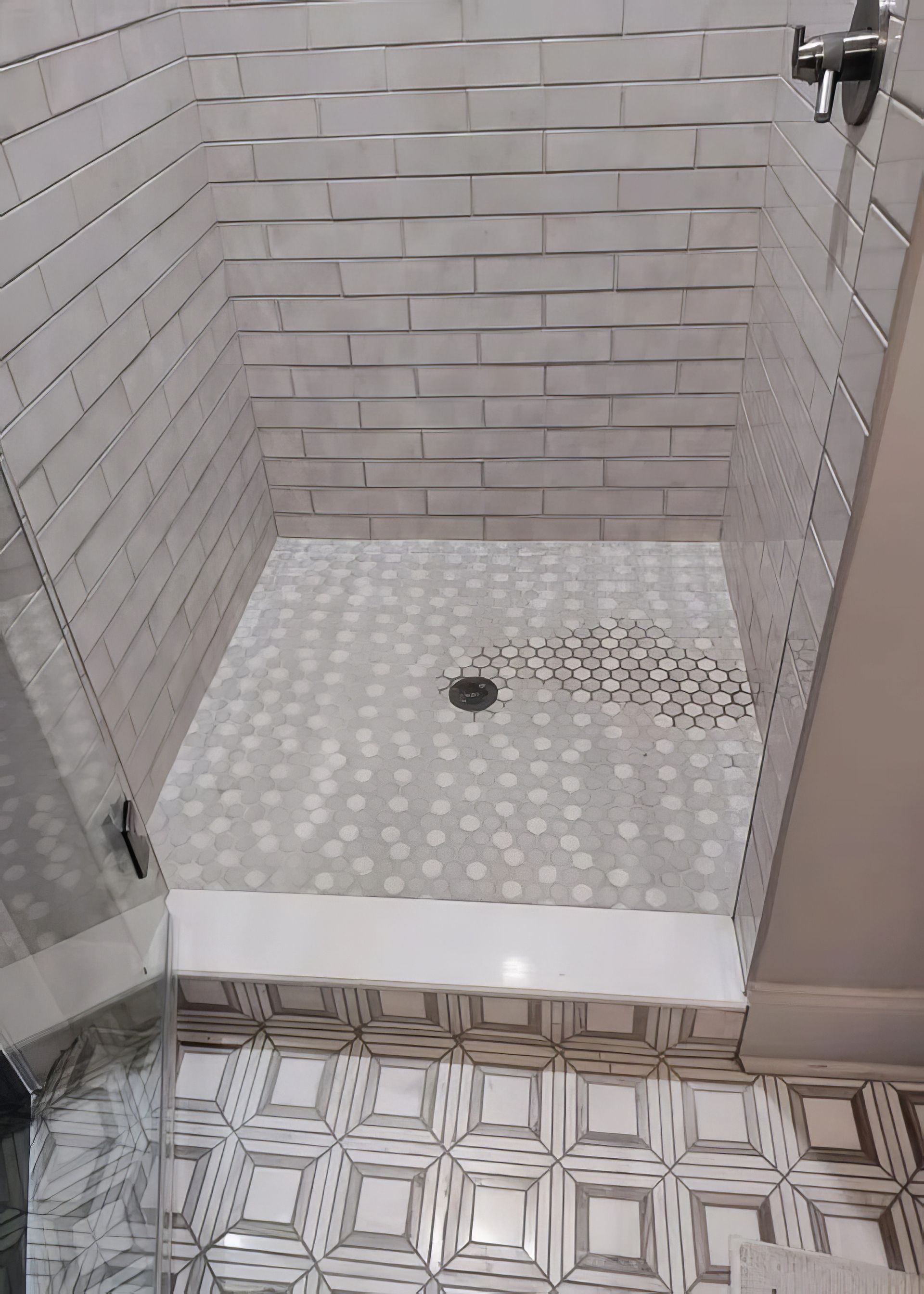 Shower stall with white brick-like tile walls and a mosaic floor. Glass door and shower head visible.
