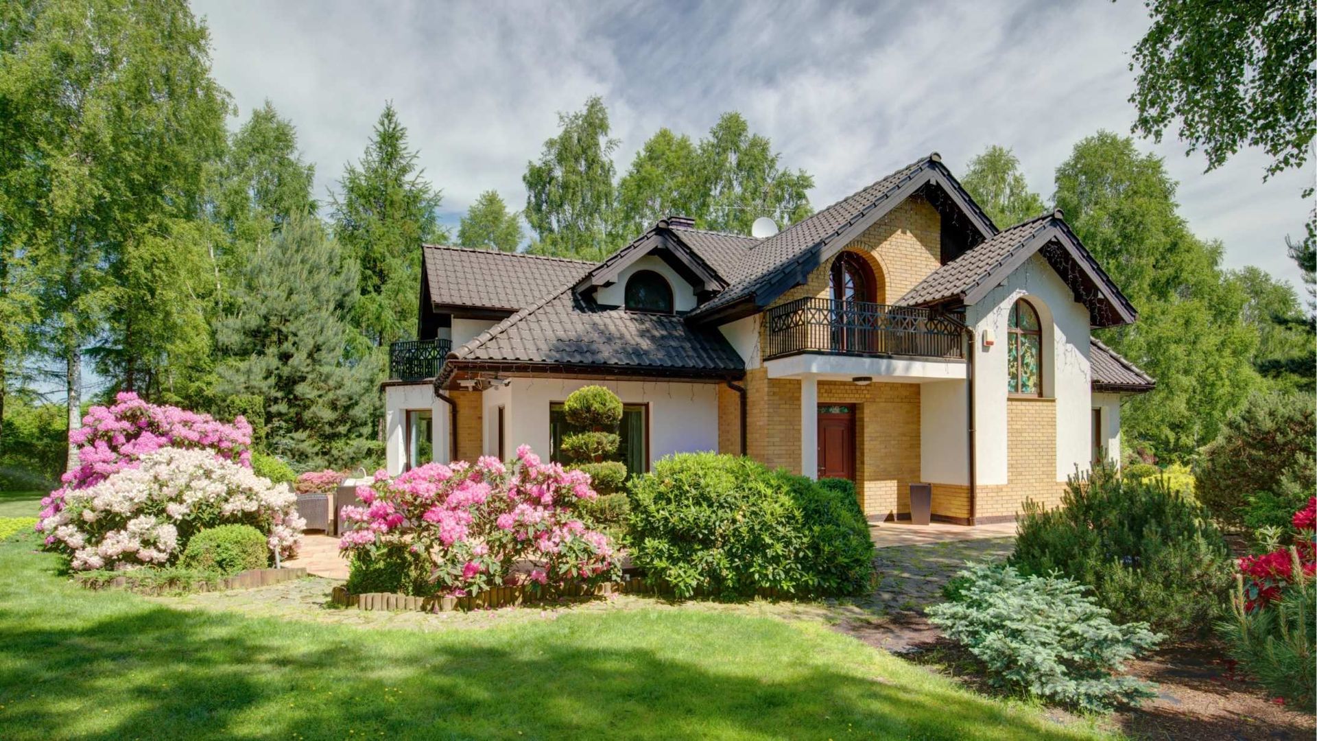 House with tan and white facade, brown roof, and lush green landscaping under a partly cloudy sky.