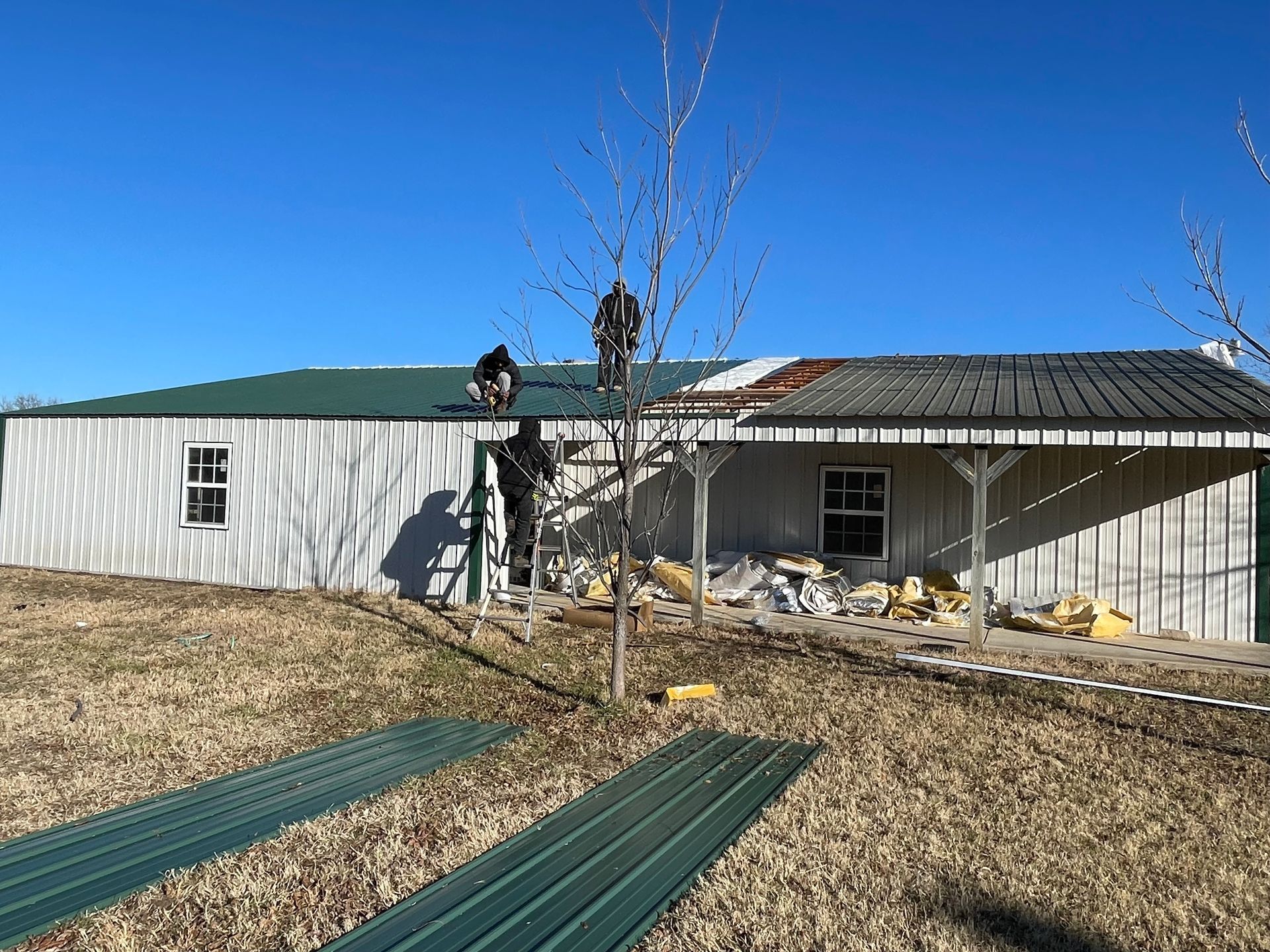Roofers replacing a metal roof on a white building. One person on the roof, another on a ladder.