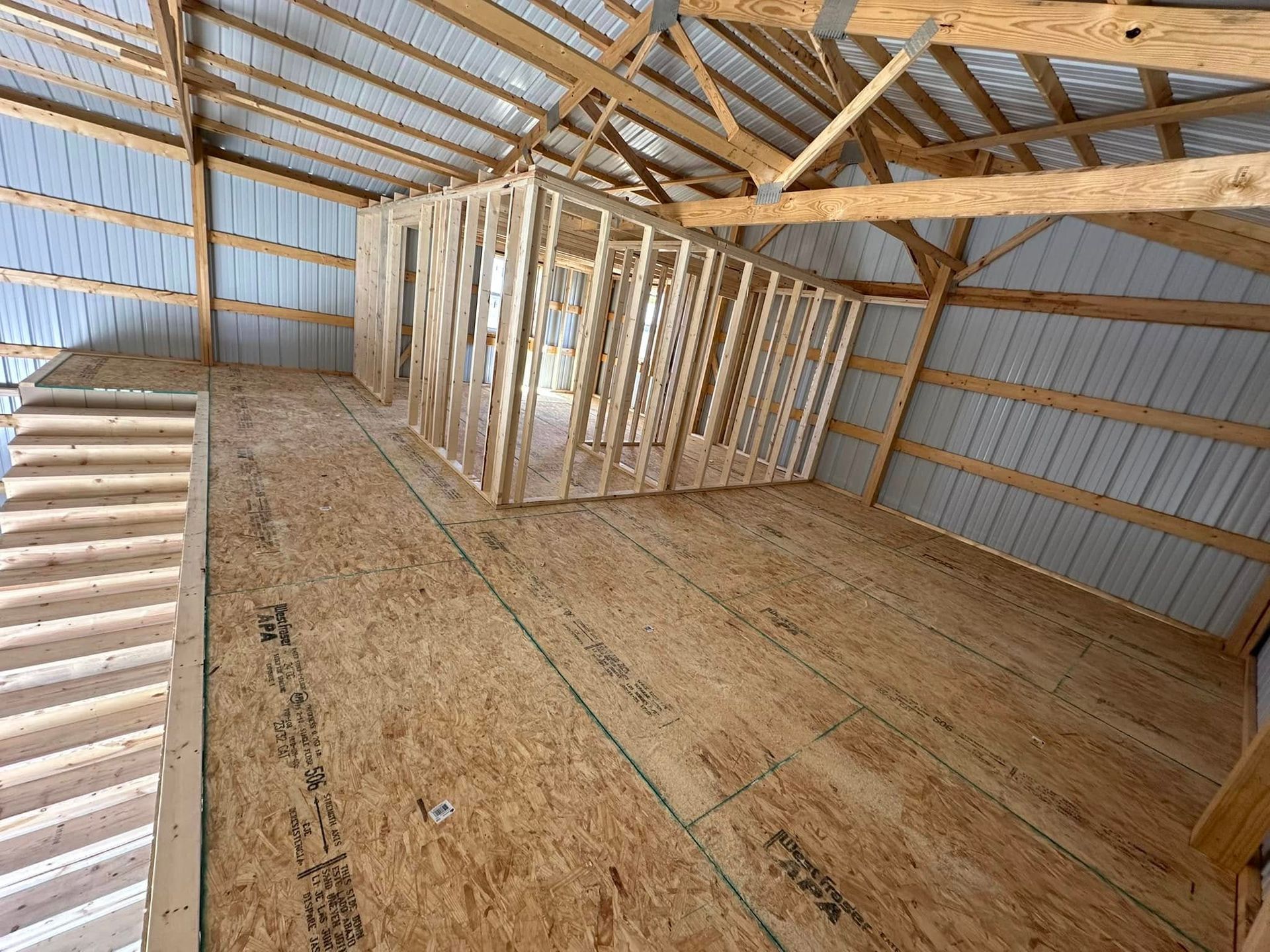 Interior view of a barn under construction with a wooden frame and exposed metal roof.