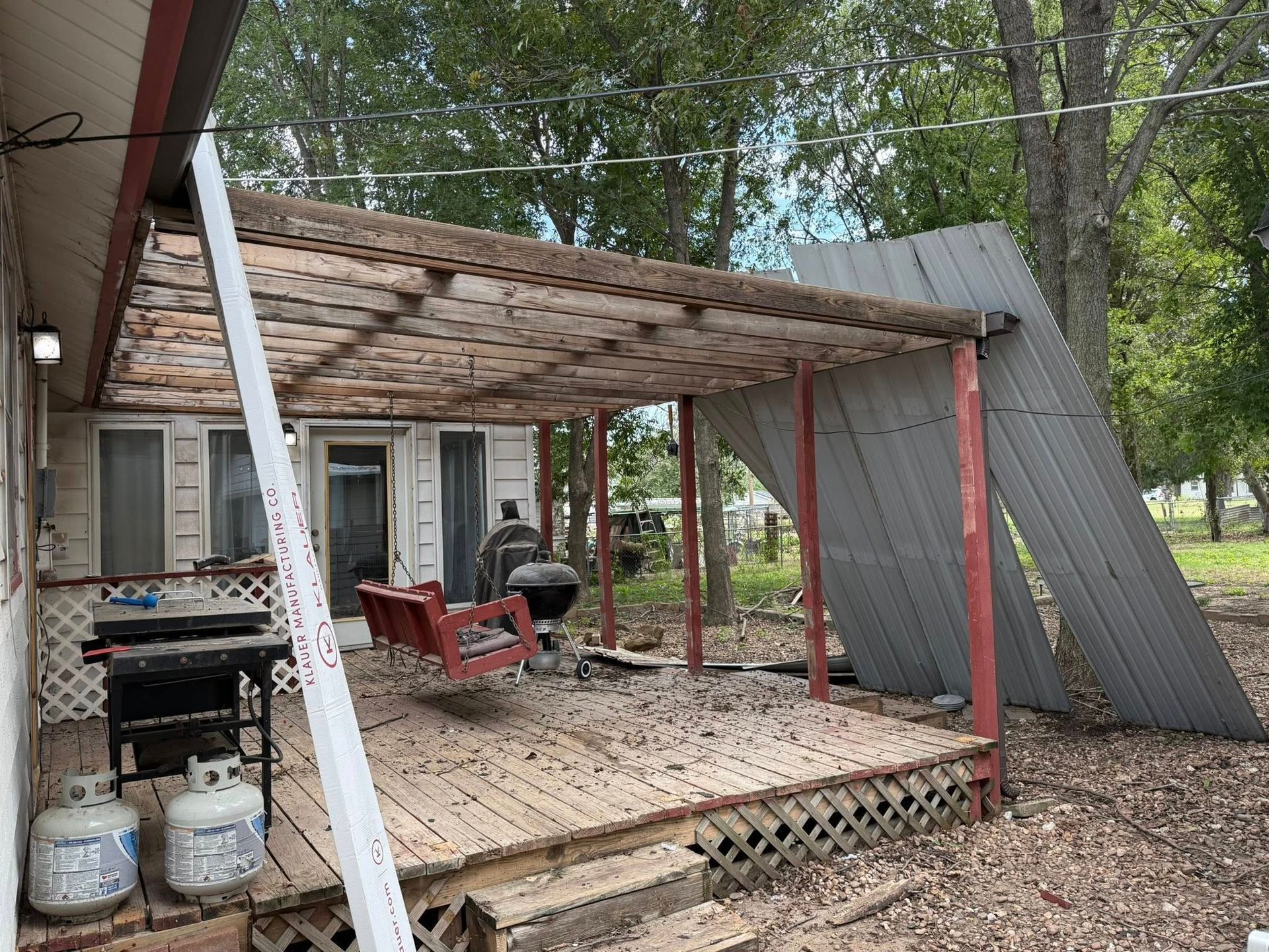 Damaged porch with a red swing, grill, and leaning metal roof in an outdoor setting.