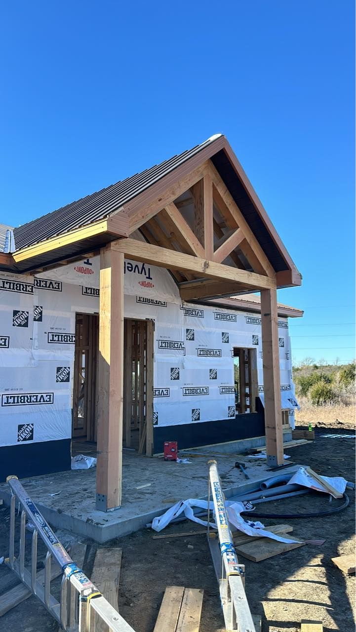 Construction of a house's entryway. Wooden beams support a roof with black shingles.