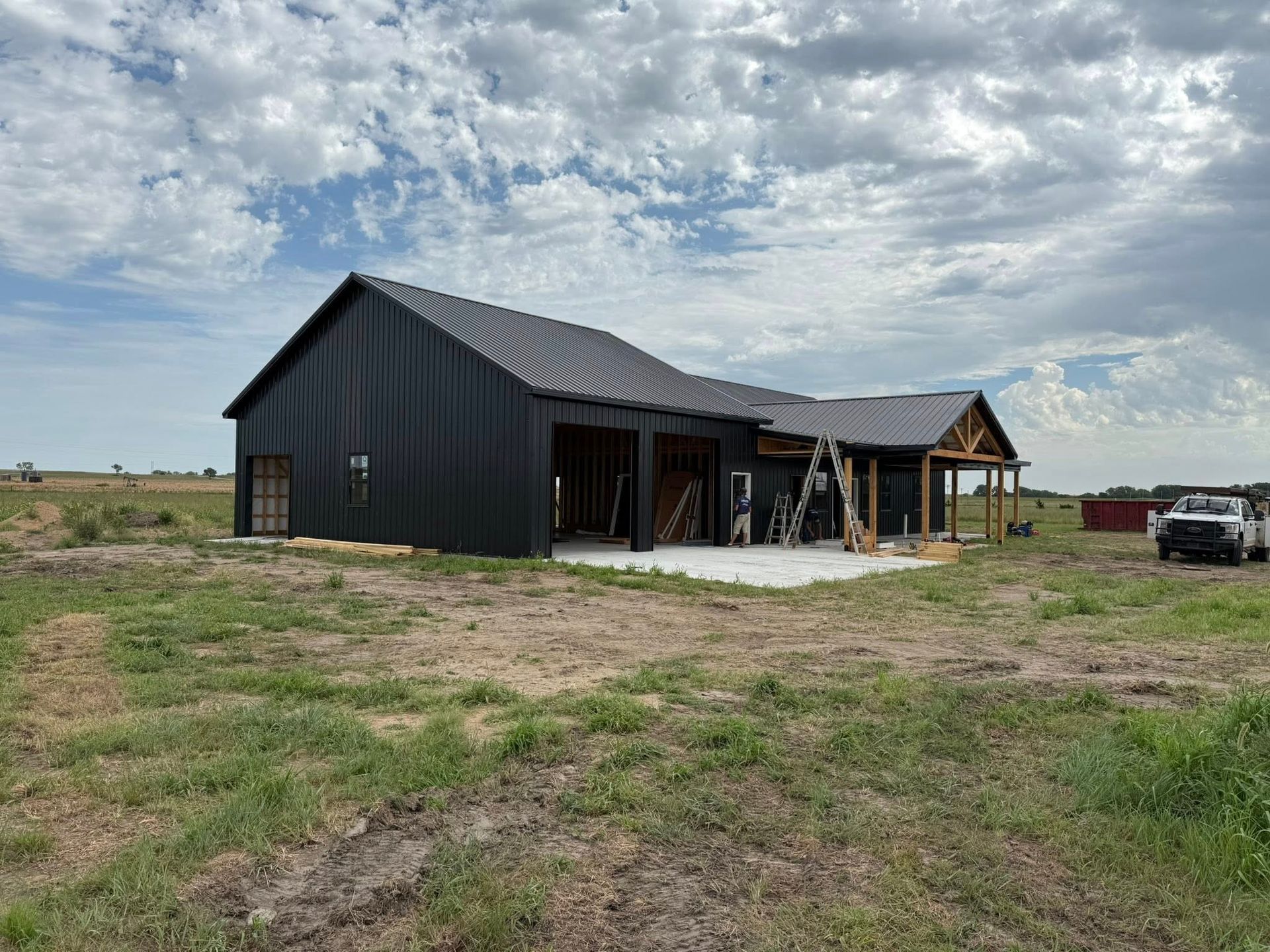 Black barn-style house under construction with a metal roof. Concrete patio and open garage. Cloudy sky.