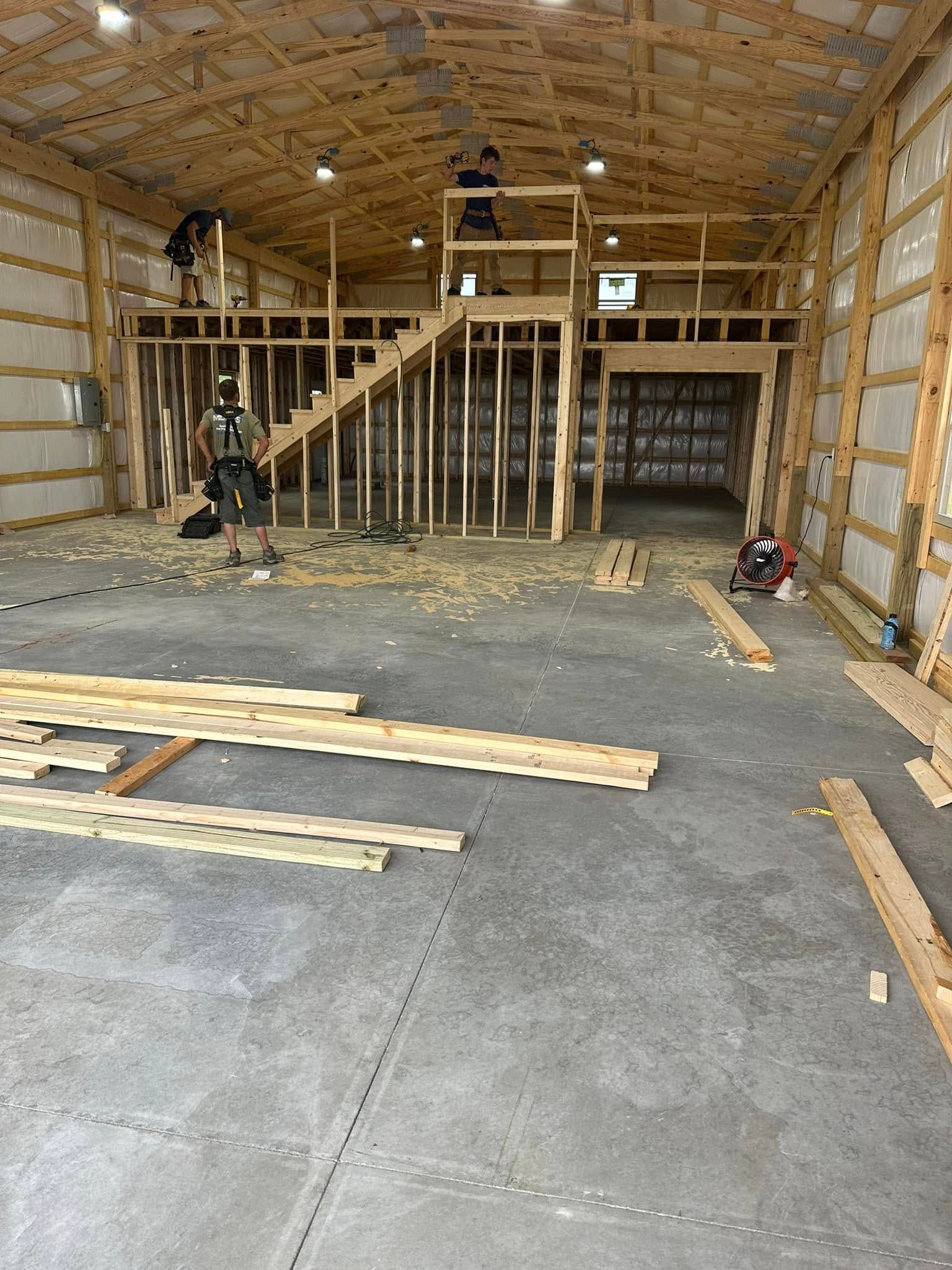 Interior view of a building under construction. Framing, stairs, and workers are visible.