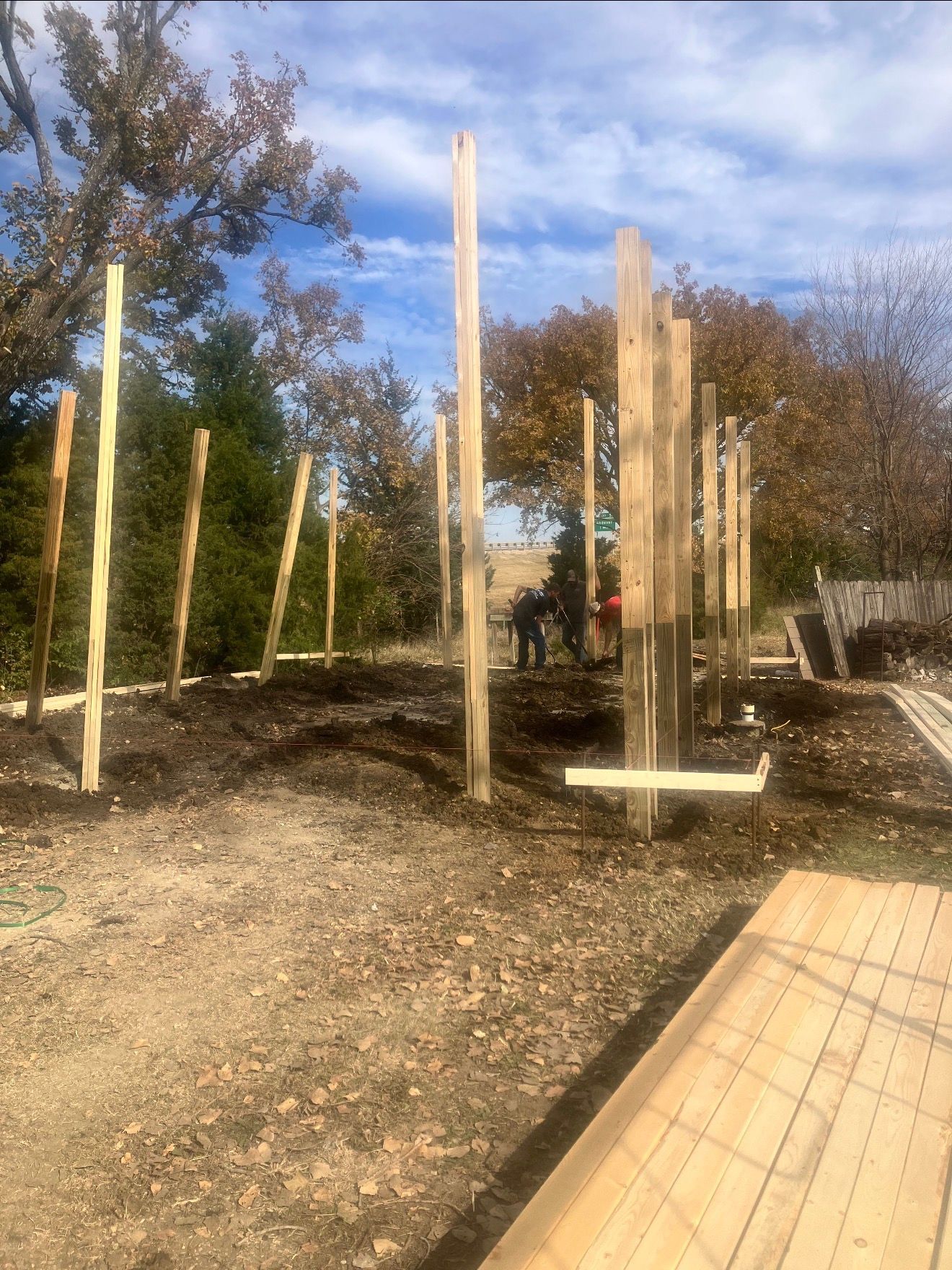 Construction site: wooden posts set in the ground, workers in the background, lumber nearby, outdoors on a cloudy day.