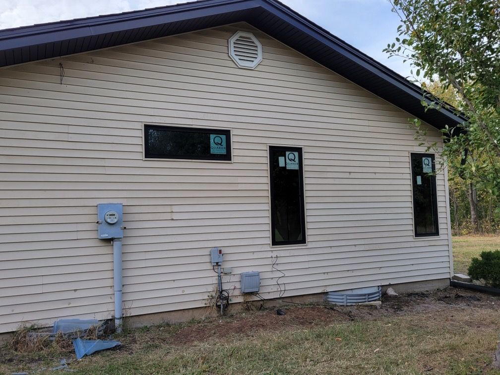 Tan-sided house with black-framed windows and a circular vent under the dark roof. Utility boxes and grass in front.