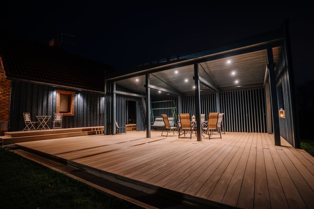 Night view of a modern deck with a covered seating area; wooden planks, lit by overhead lights.